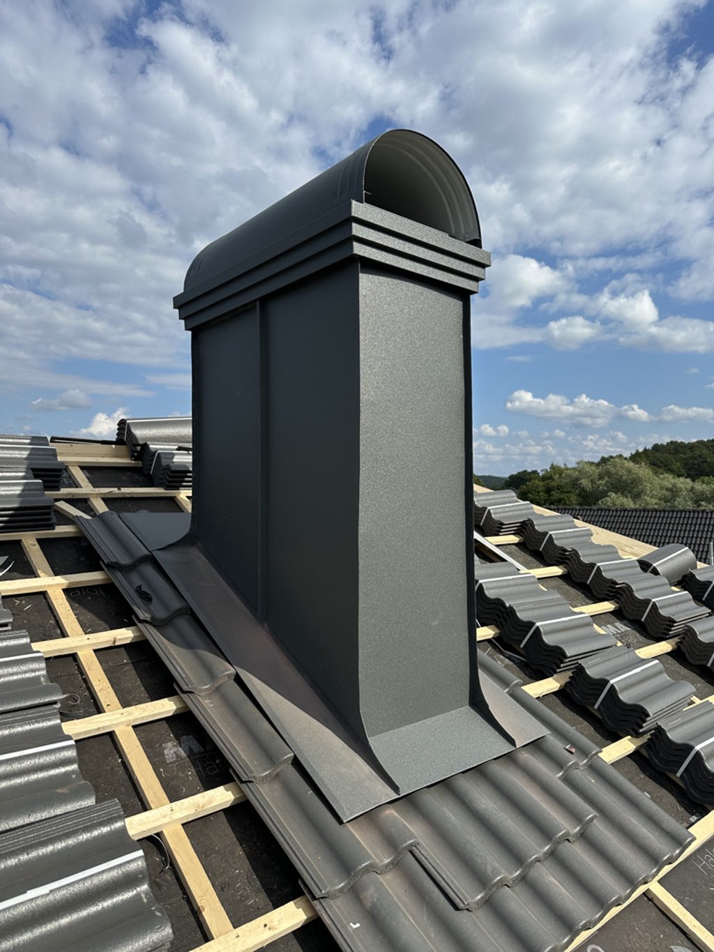 En takslev med en metallskorsten på, omgiven av takpannor på ett tak under en molnig himmel.