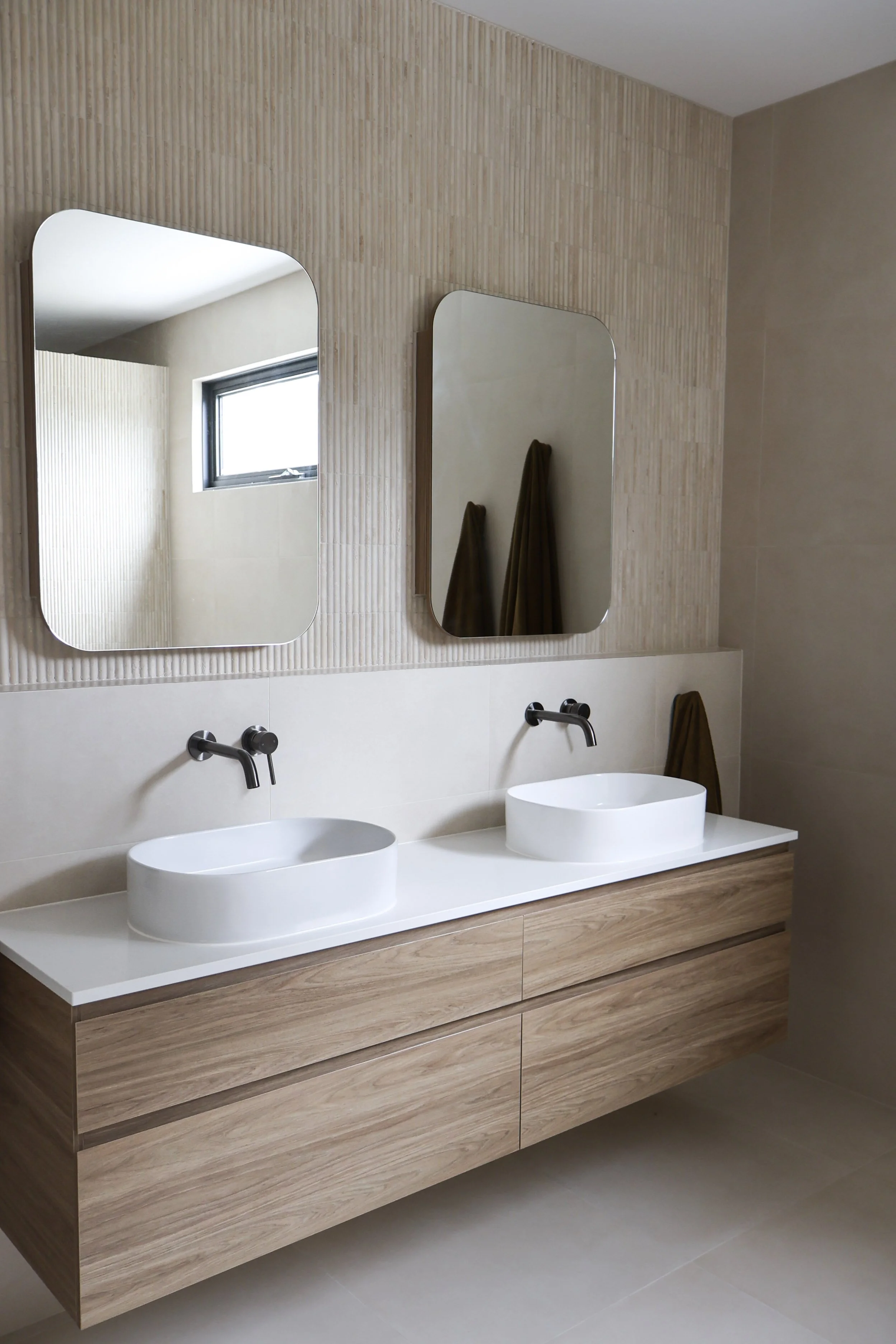 Modern bathroom with double white vessel sinks, wall-mounted faucets, and wooden vanity. Two rectangular mirrors above sinks reflect a small window and towels hanging on the wall.