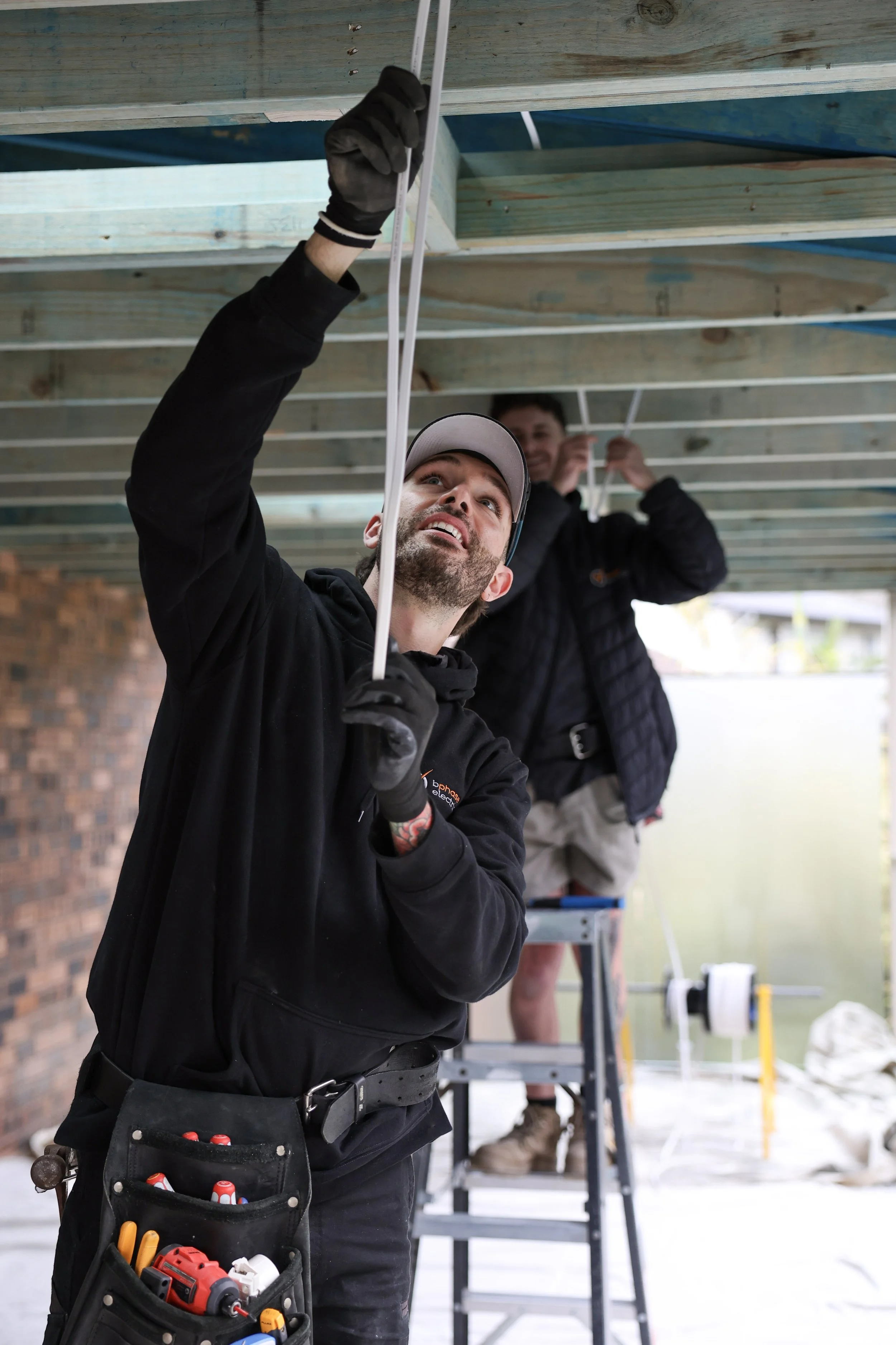 Two workers installing electrical wiring under a house, with one on a ladder and the other reaching up, both wearing tools belts.