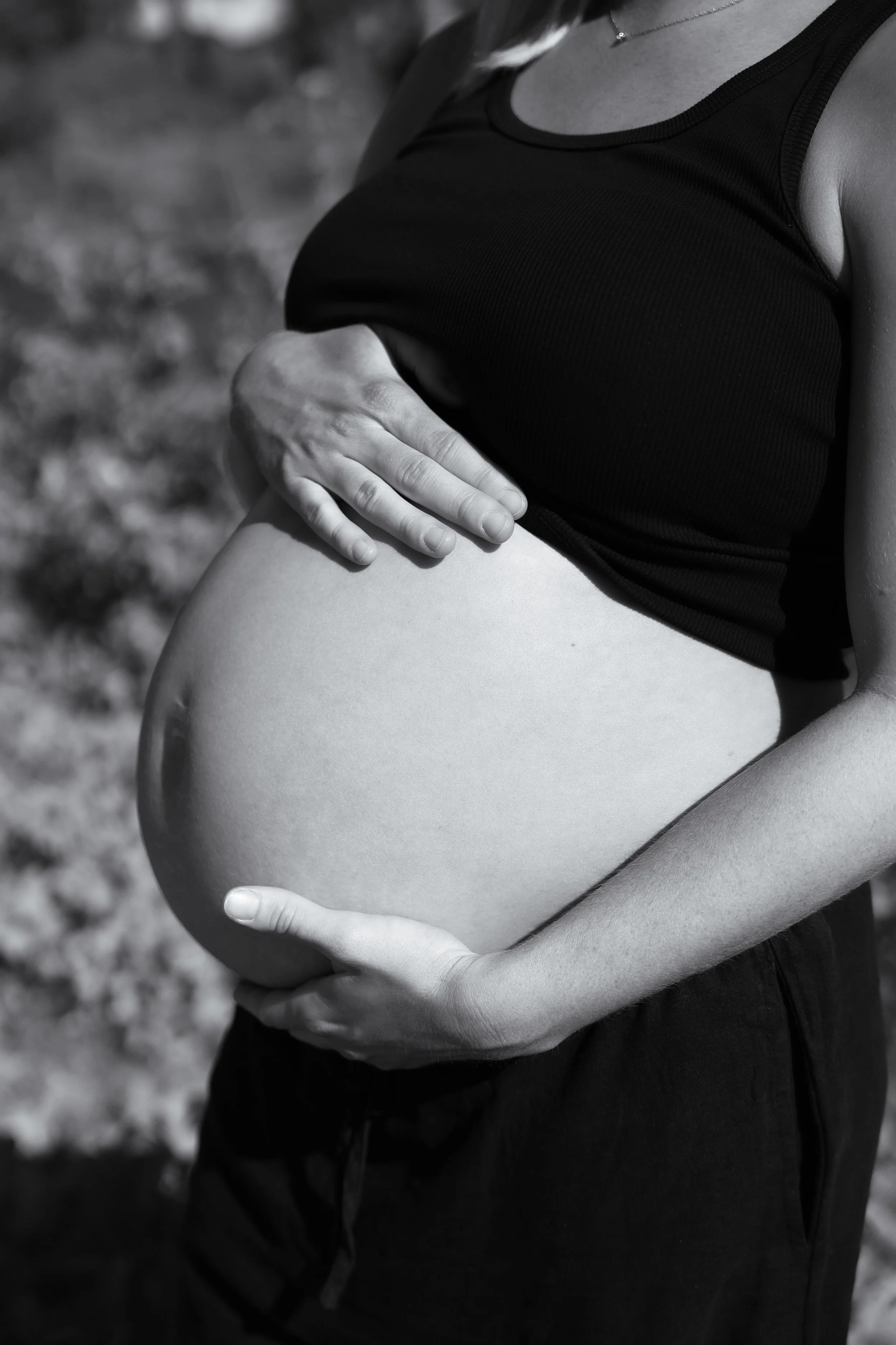 A pregnant woman in a black tank top cradling her belly with both hands, outdoors.