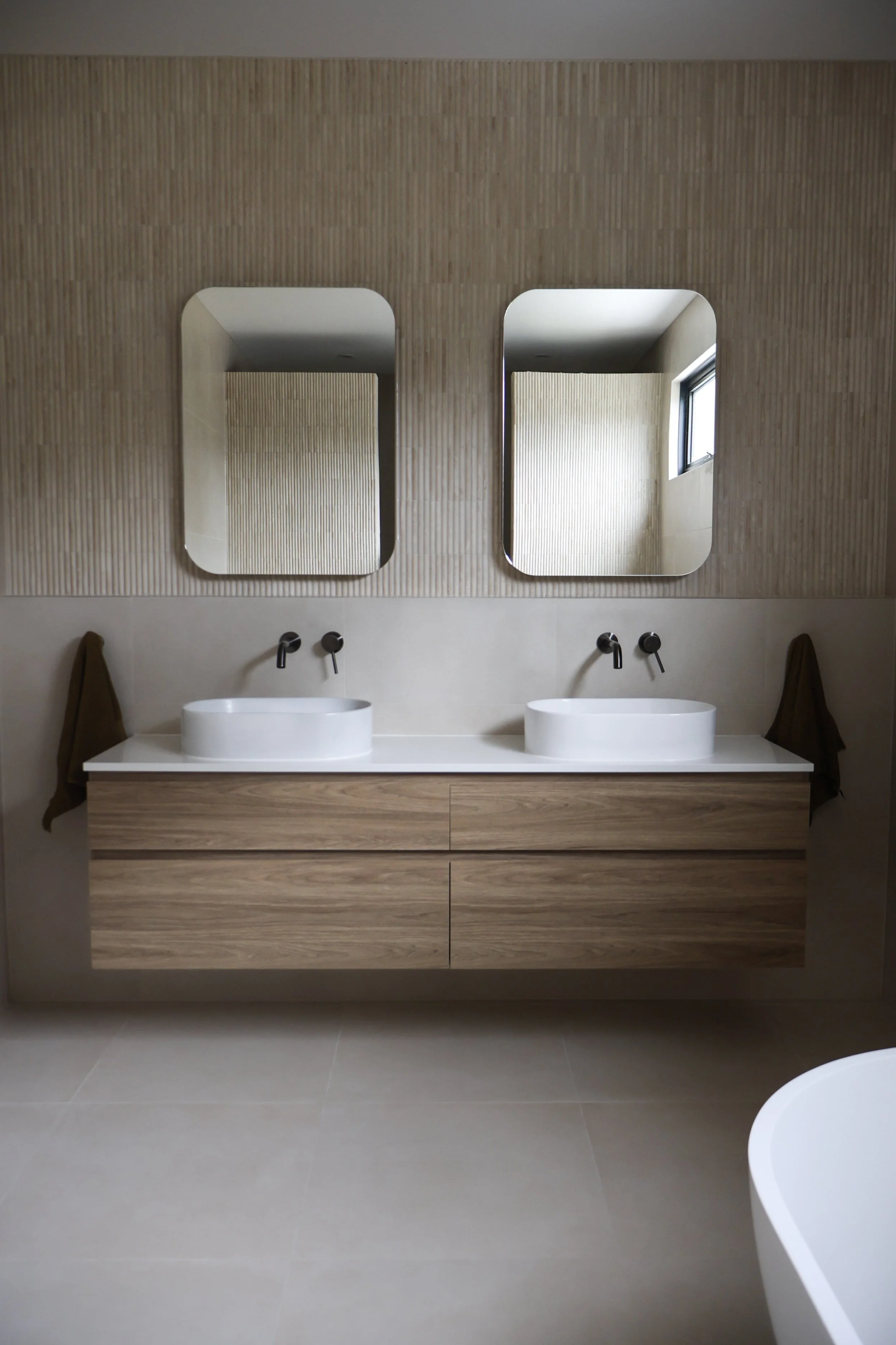 Modern bathroom with two oval vessel sinks on a wooden vanity, two mirrors above, and a small window reflected in the right mirror.