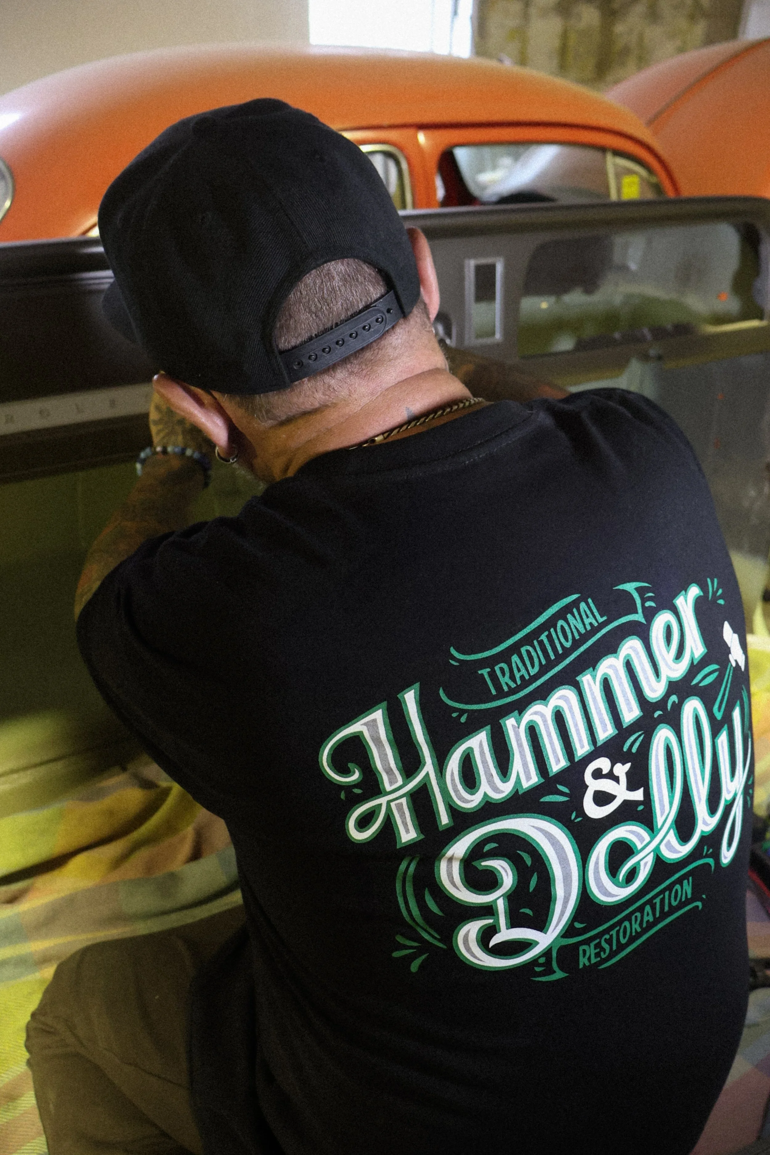 A person wearing a black baseball cap and black T-shirt with 'Traditional Hammer & Dolly Restorations' written on it, working on restoring a vintage vehicle inside a garage, with other vintage cars visible in the background.