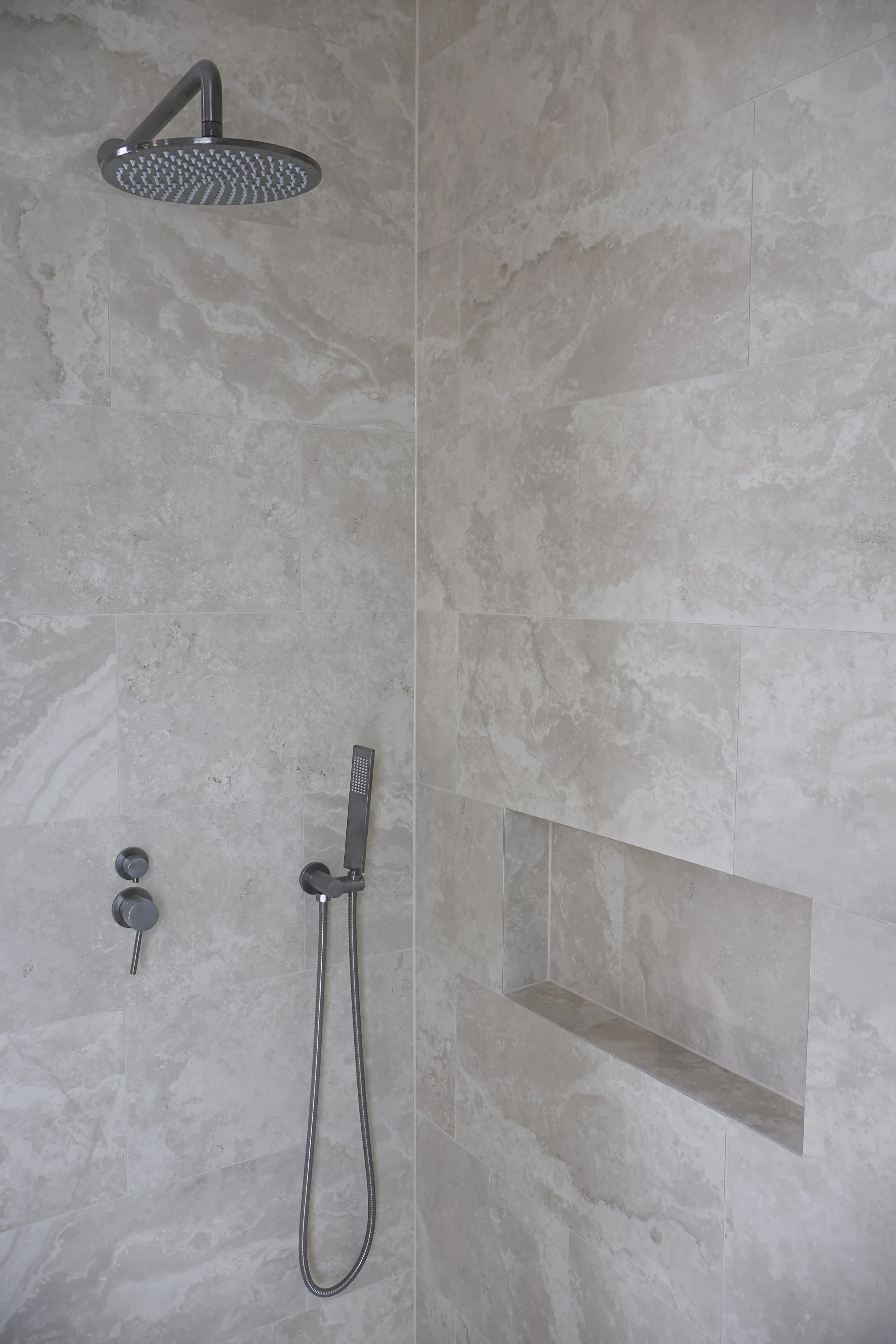 A modern shower with a round rainfall showerhead, a handheld showerhead, and beige tiled walls with a built-in niche for toiletries.