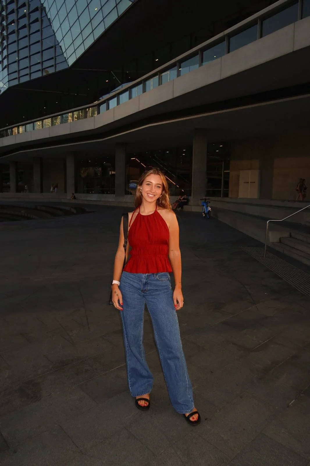 A young woman stands outdoors in front of a modern building with glass windows, wearing a red sleeveless top, blue jeans, and black sandals, smiling at the camera during what appears to be sunset or evening.