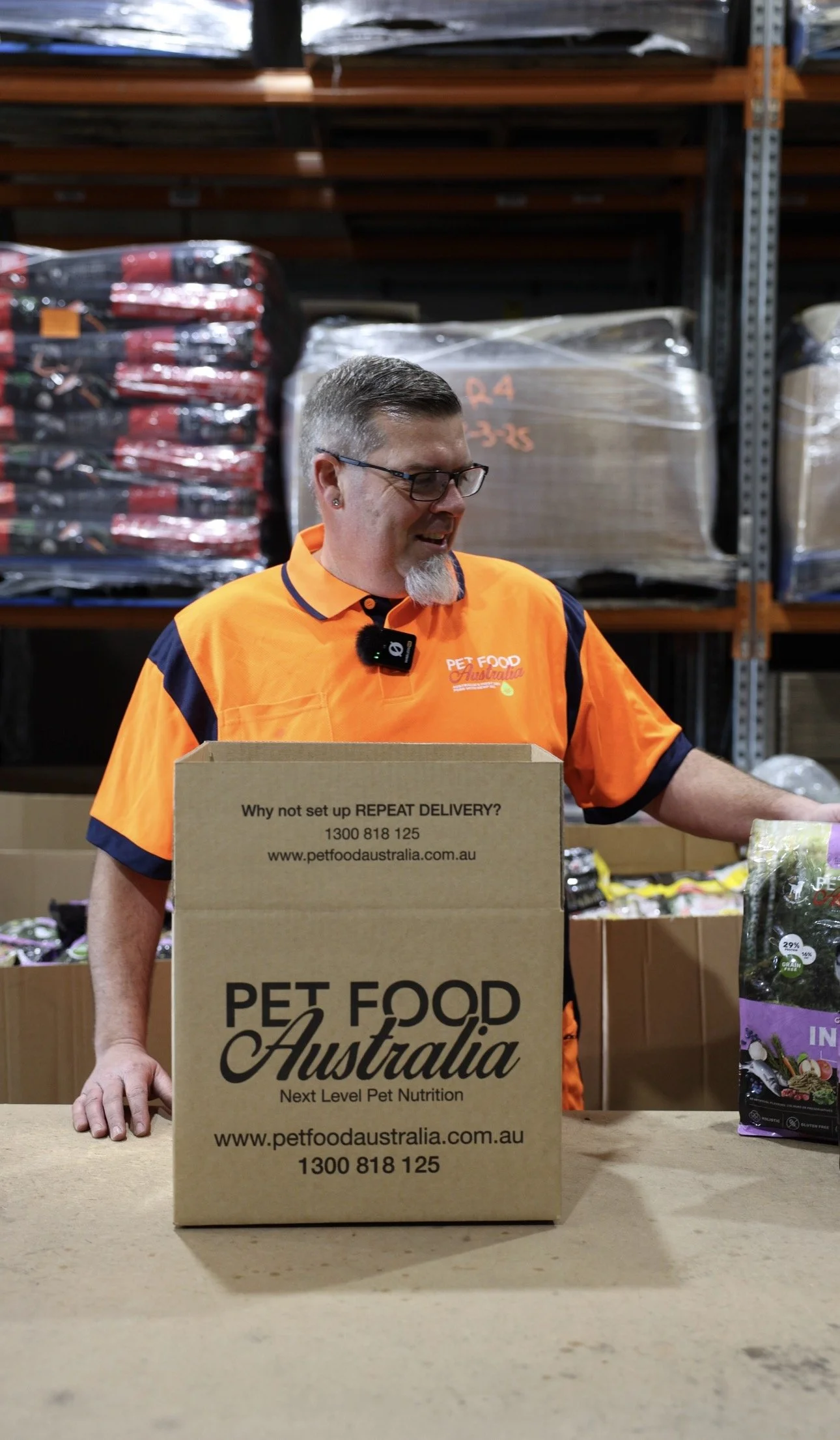 A man in an orange Pet Food Australia uniform stands behind a table with a large cardboard box displaying the company's logo and contact information, with shelves of pet food bags in the background.