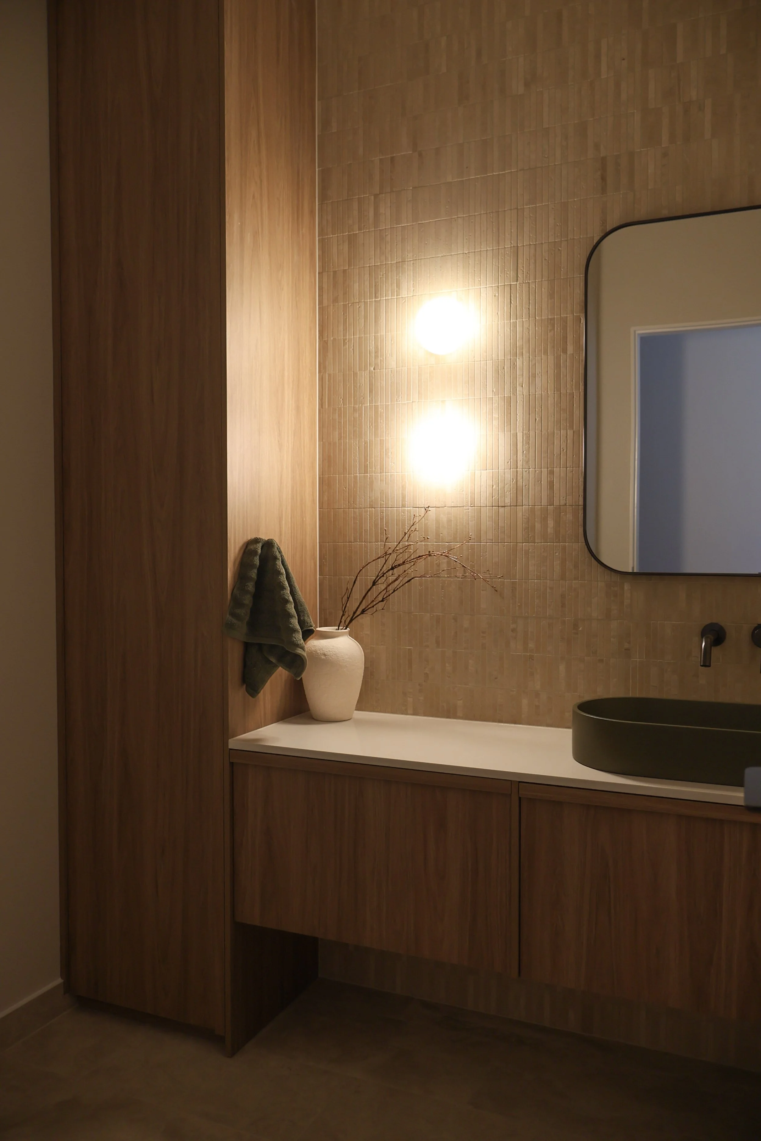 Modern bathroom vanity area with wooden cabinet, white countertop, green vessel sink, large mirror, wall-mounted faucet, warm lighting, beige tiled wall, decorative vase with branches, gray towel hanging on the side.