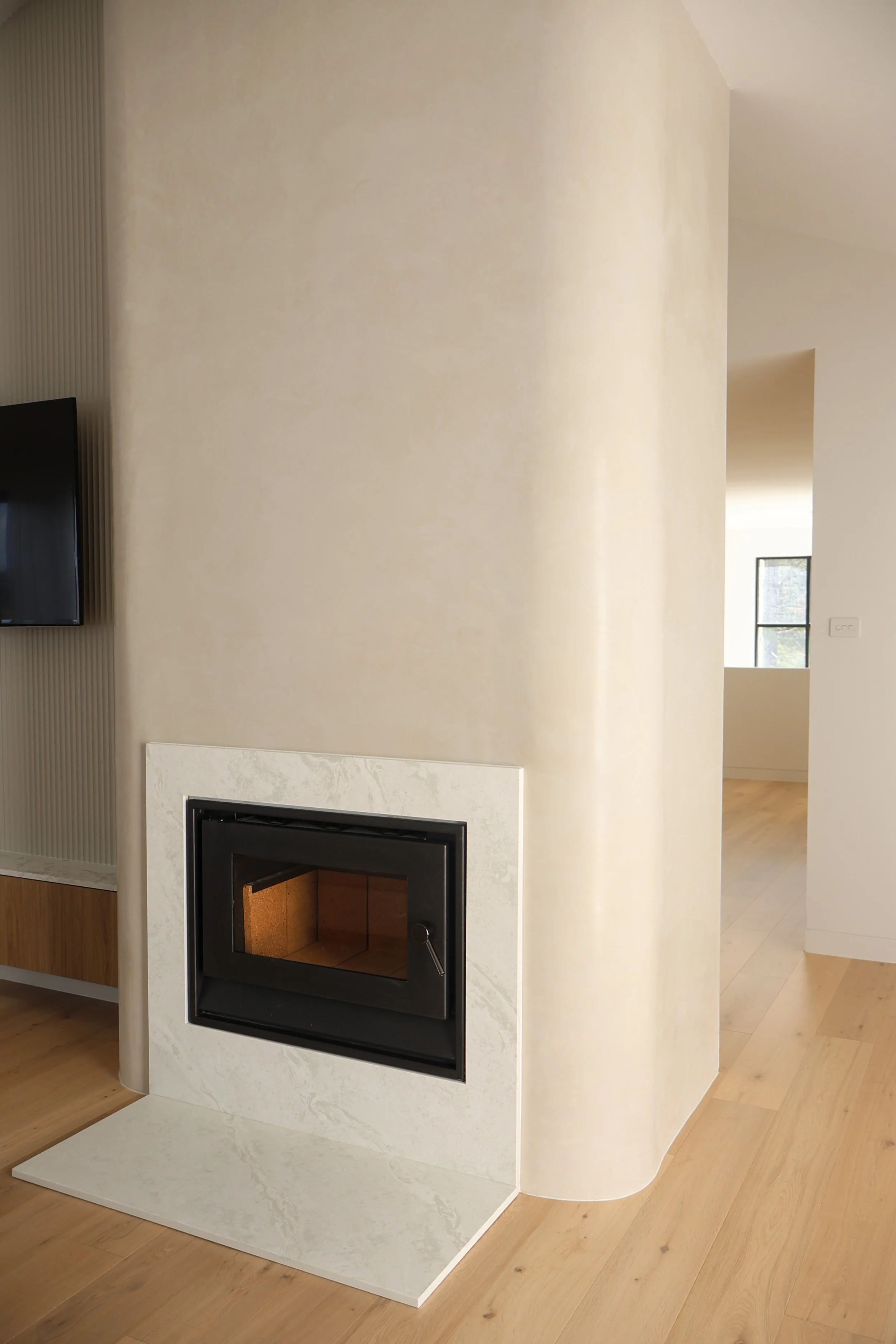 Modern indoor fireplace with a black metal insert, beige marble surround, curved wall, and light wooden floor.