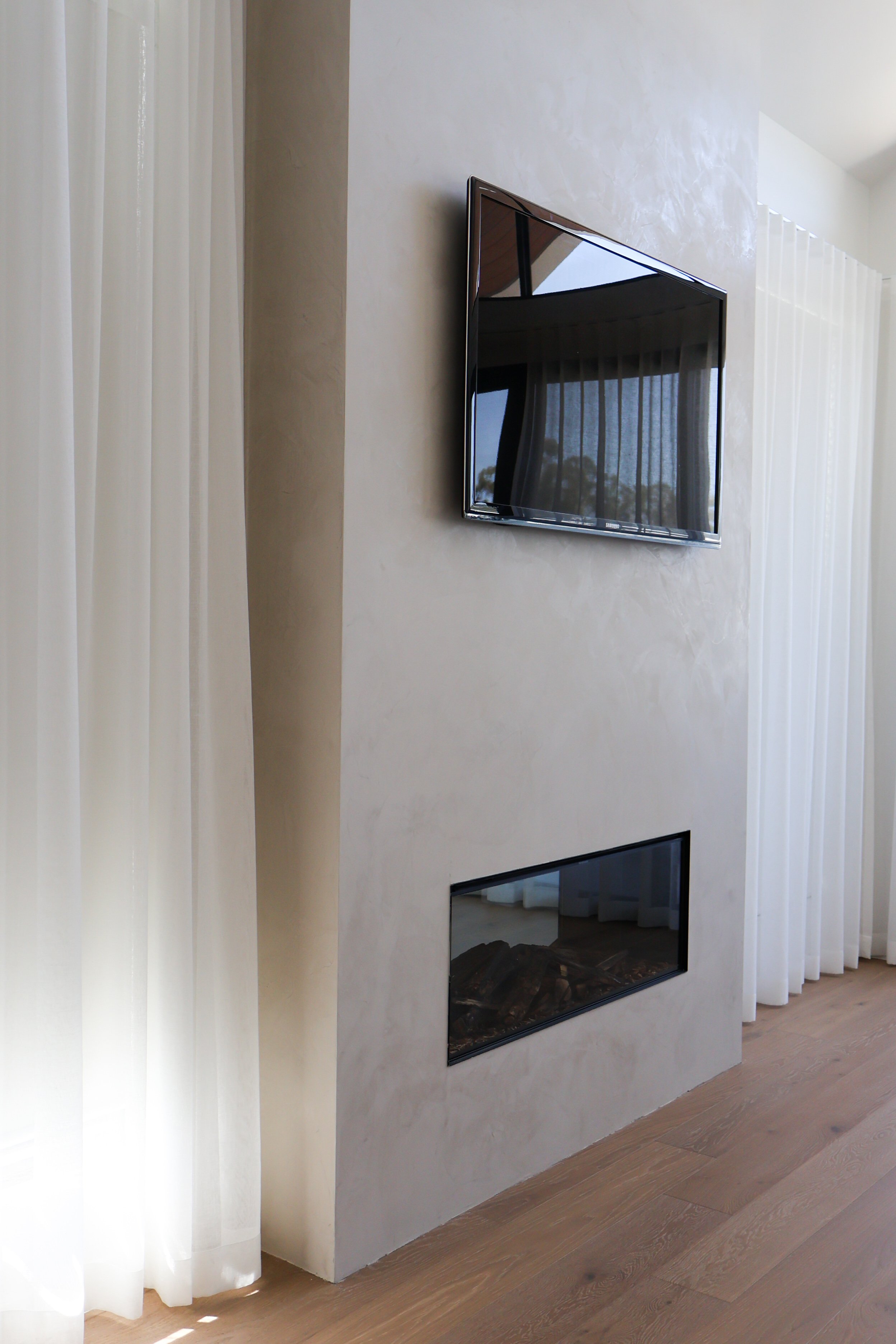 A modern living room with a wall-mounted flat-screen TV and a rectangular fireplace below it, with white curtains on the sides and wooden flooring.