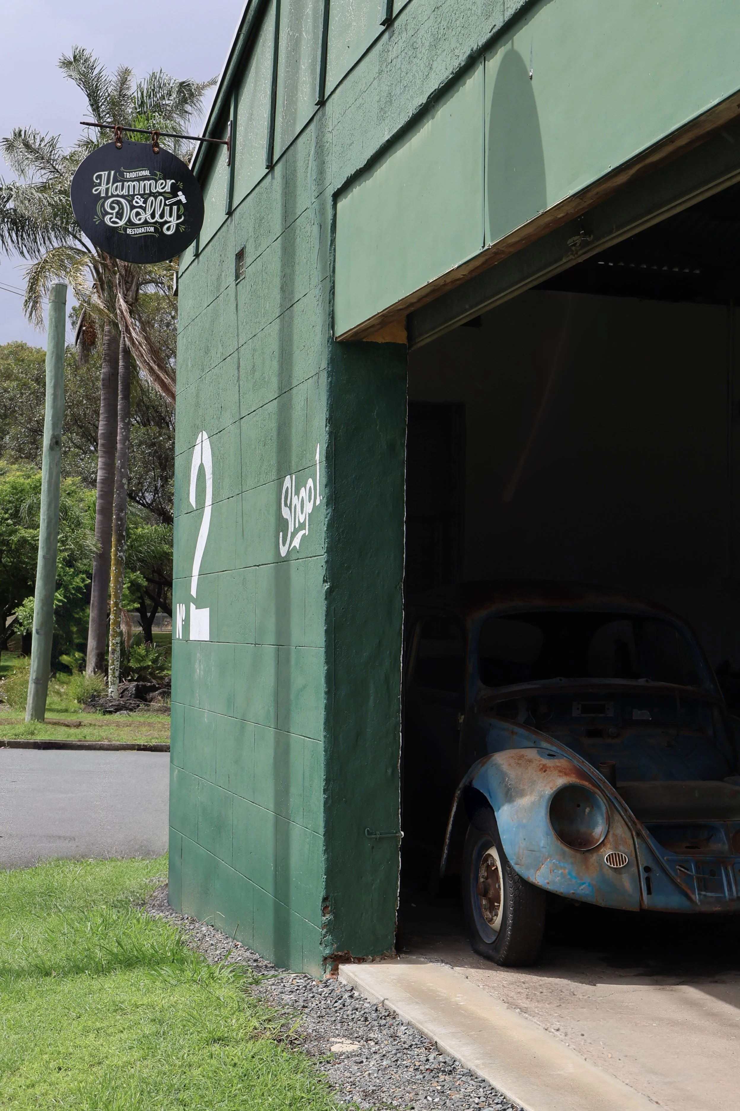 A green building with a sign that reads 'Hammer & Dolly' outside. The wall has white text and a question mark with the words 'Shop No 2'. An old, rusted blue Volkswagen Beetle is in a garage area partially inside the building, with some trees and pal