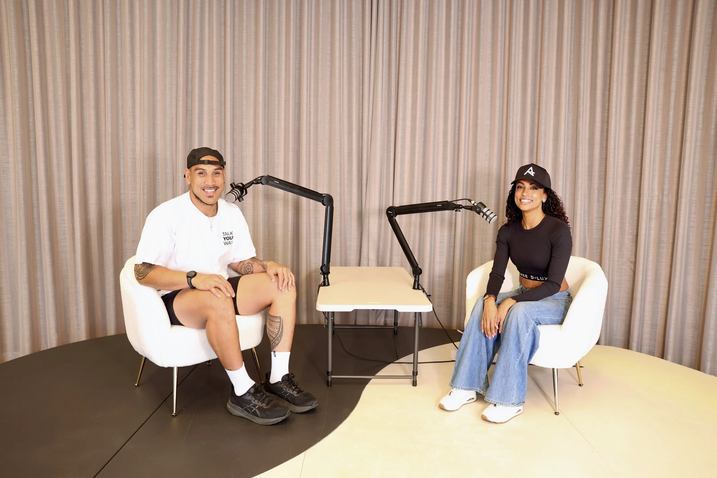 Two people sitting in white chairs with microphones on a small table between them, in front of a beige curtain backdrop.