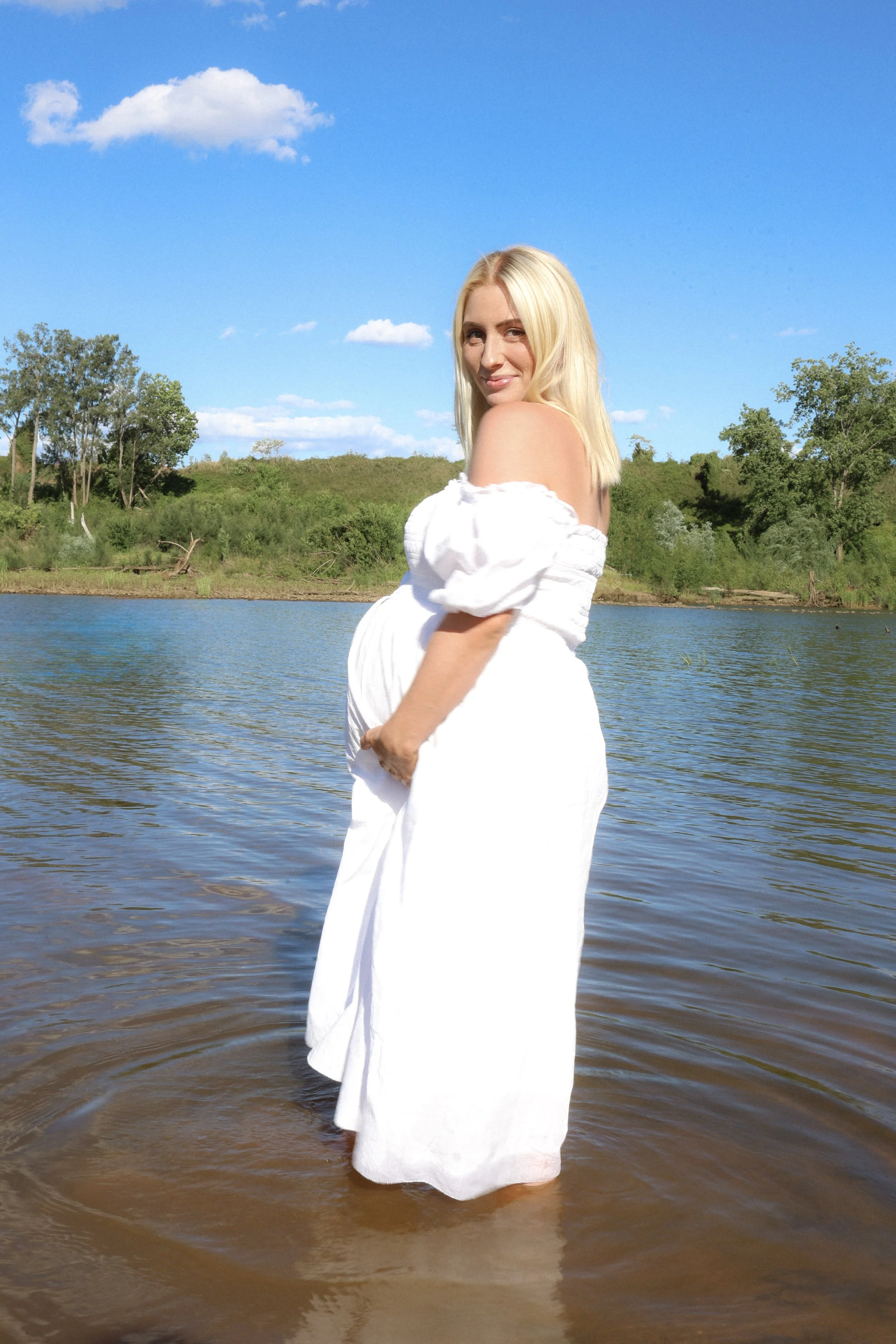 A smiling blonde woman in a white off-shoulder dress standing in a river on a sunny day with green trees, grass, and a blue sky with a few clouds in the background.