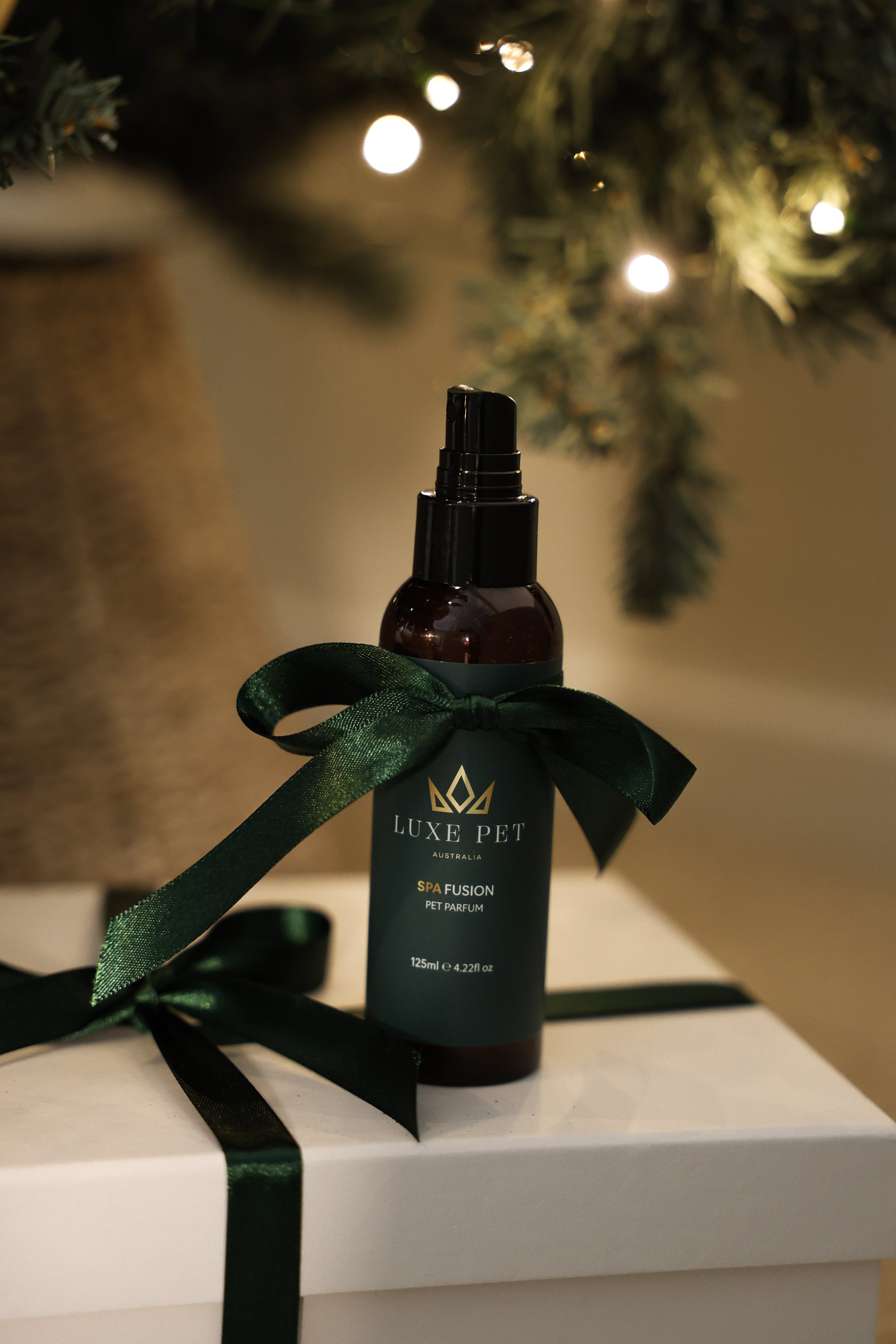 Gift box with a bottle of Luxe Pet spa perfume, decorated with a dark green ribbon, placed under a Christmas tree with lights.