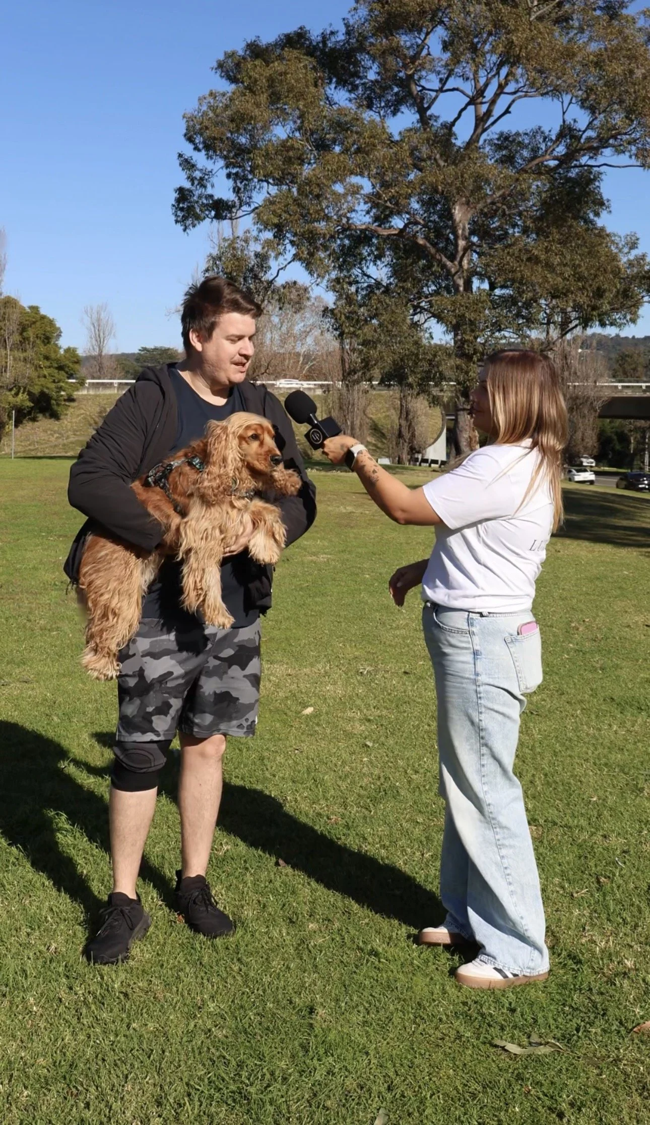 A man holding a golden retriever being interviewed by a woman with a microphone in a park.