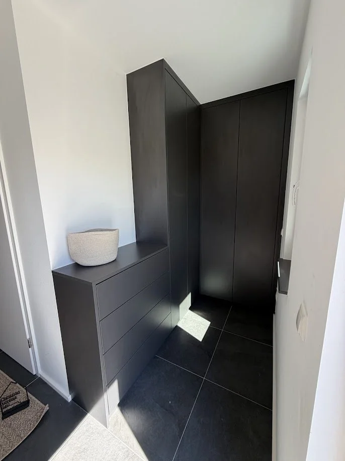Ein kleiner Eingangsbereich mit schwarzem Schrank und Kommode, eine weiße dekorative Schüssel auf der Kommode, dunkle Fliesen an der Fußboden, weißer Wand, natürliche Tageslichtbeleuchtung.