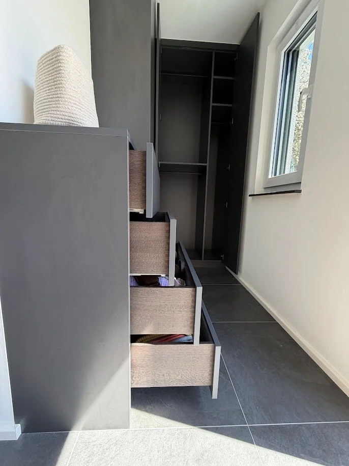 Leeres schwarzes Kleiderschrank mit offenen Schubladen vor einem Fenster in einem weißen Raum mit dunklem Fliesenboden.