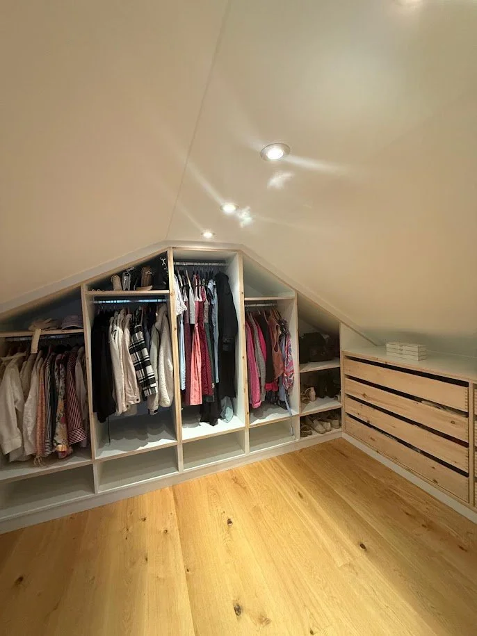 Kleiderschrank mit Kleidung in einem Dachboden, helle Holzböden, weiße Regale, teilweise kühl beleuchtet.