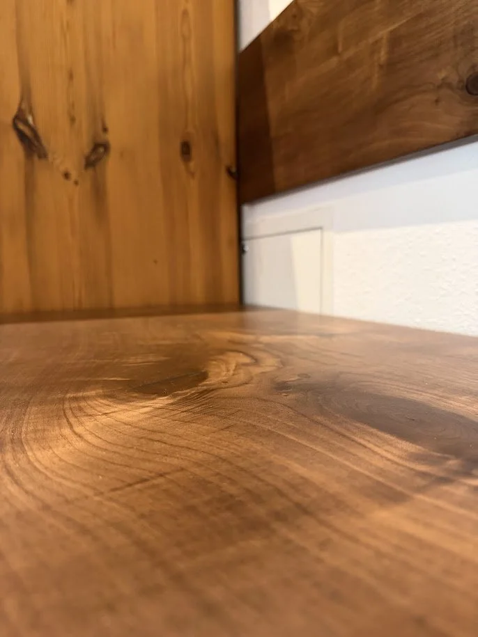 Nahaufnahme einer Holzoberfläche mit sichtbaren Maserungen, im Hintergrund eine weiße Wand und eine Holzvertäfelung.