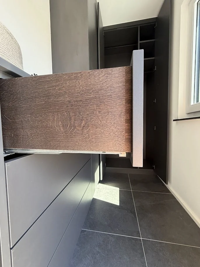 Ein offener Schrank mit mehreren Fächern und eine sichtbare Fensterseite in einem Raum mit dunklem Fliesenboden.