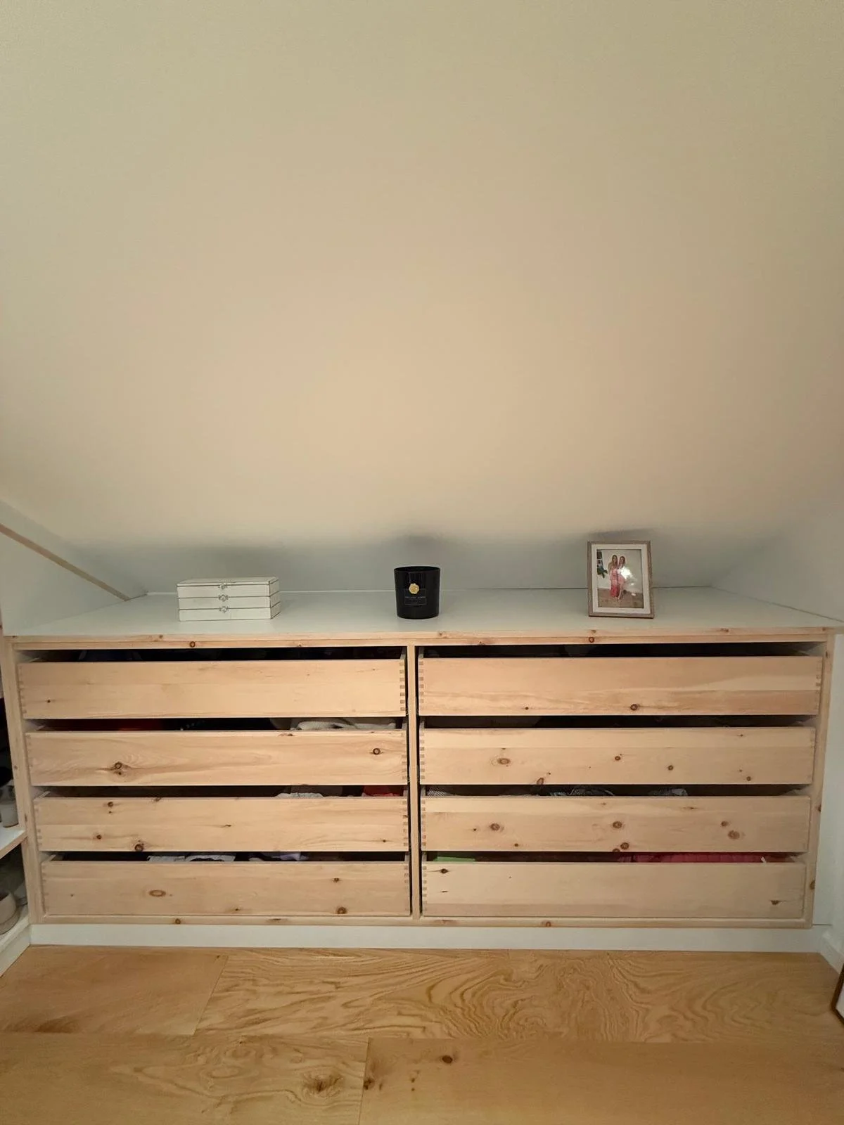 Ein Holzkommode mit acht Schubladen vor der Wand. Oben auf der Kommode liegen ein Stapel weißer Bücher, eine schwarze Vase und ein Bilderrahmen mit Foto.