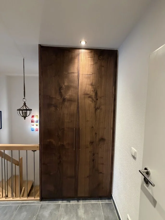 Ein kleiner Raum mit einer großen Holztür, heller Wand, einer Treppe im Hintergrund und einer Deckenleuchte.