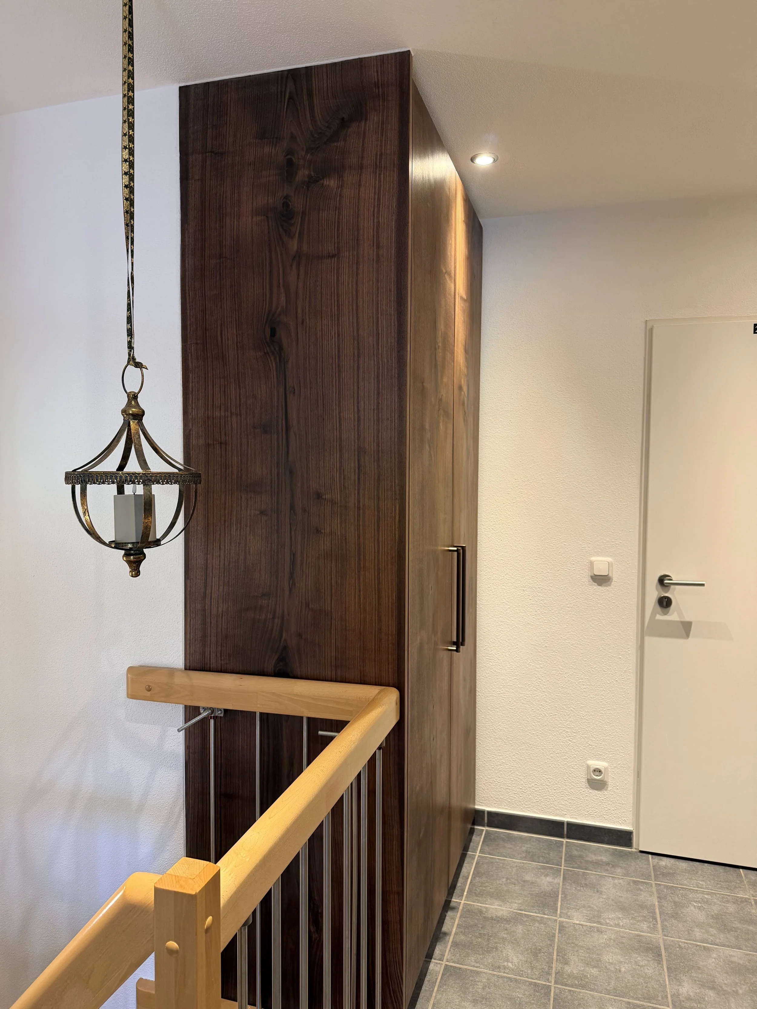 Ein Flur mit weißer Wand, einem dunklen Holz-Schrank und einem Treppengeländer aus Holz. Es gibt eine Deckenleuchte, eine Wandbuchse und eine Tür.