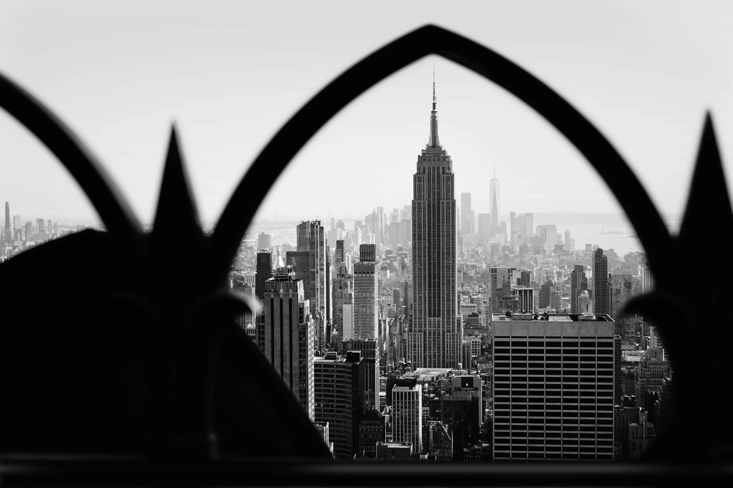 Black and white photo of the New York City skyline, with the Empire State Building prominently visible through a decorative iron fence in the foreground.