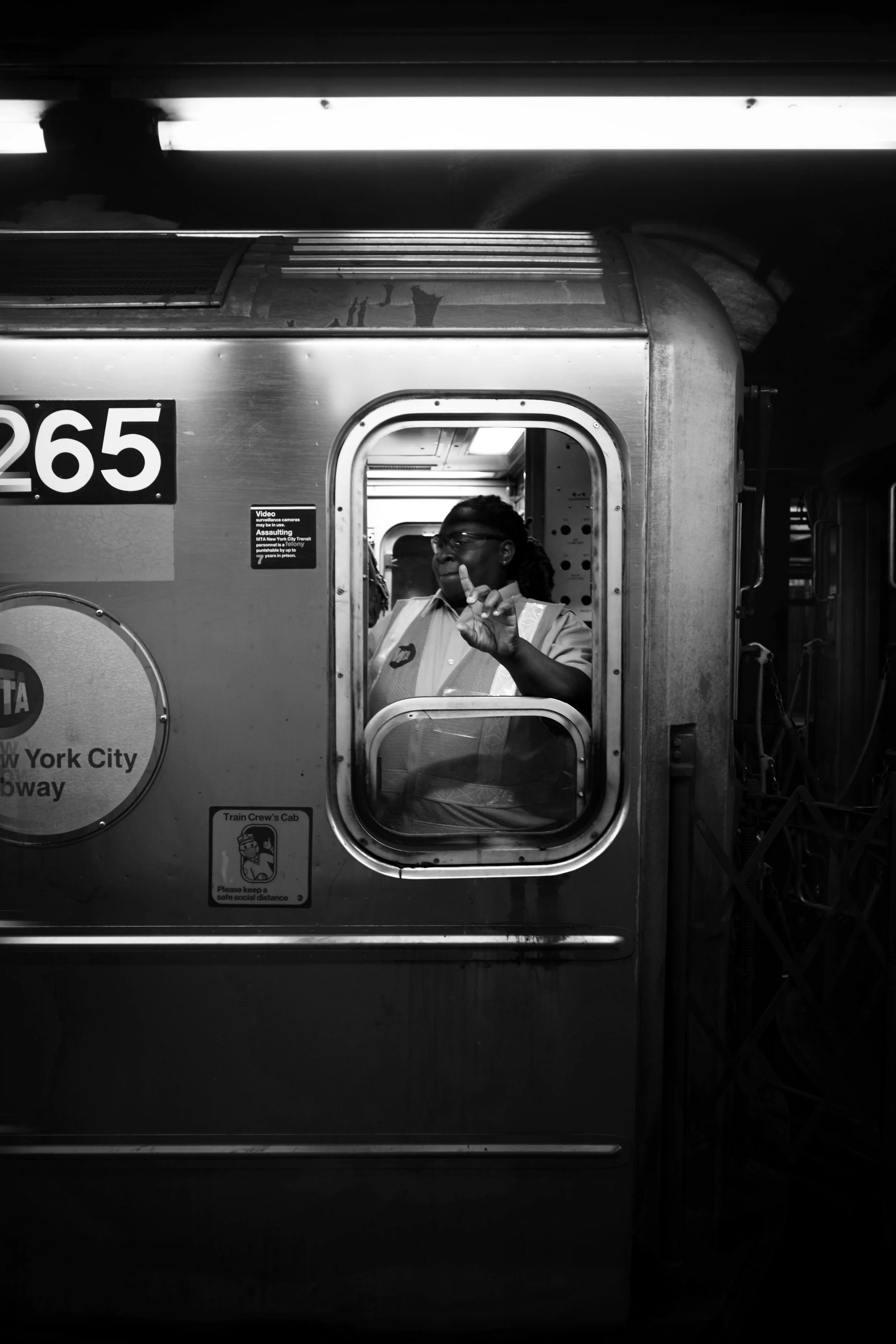A person inside a New York City subway train, visible through a window, making a peace sign gesture. The train has the identification number 265 and signs indicating it is on the NYC subway line.