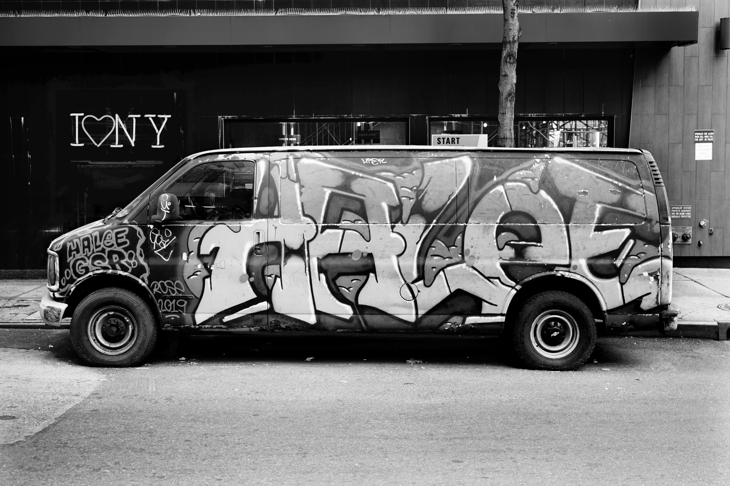 A van with graffiti art parked on a city street, with a tree and storefront in the background.