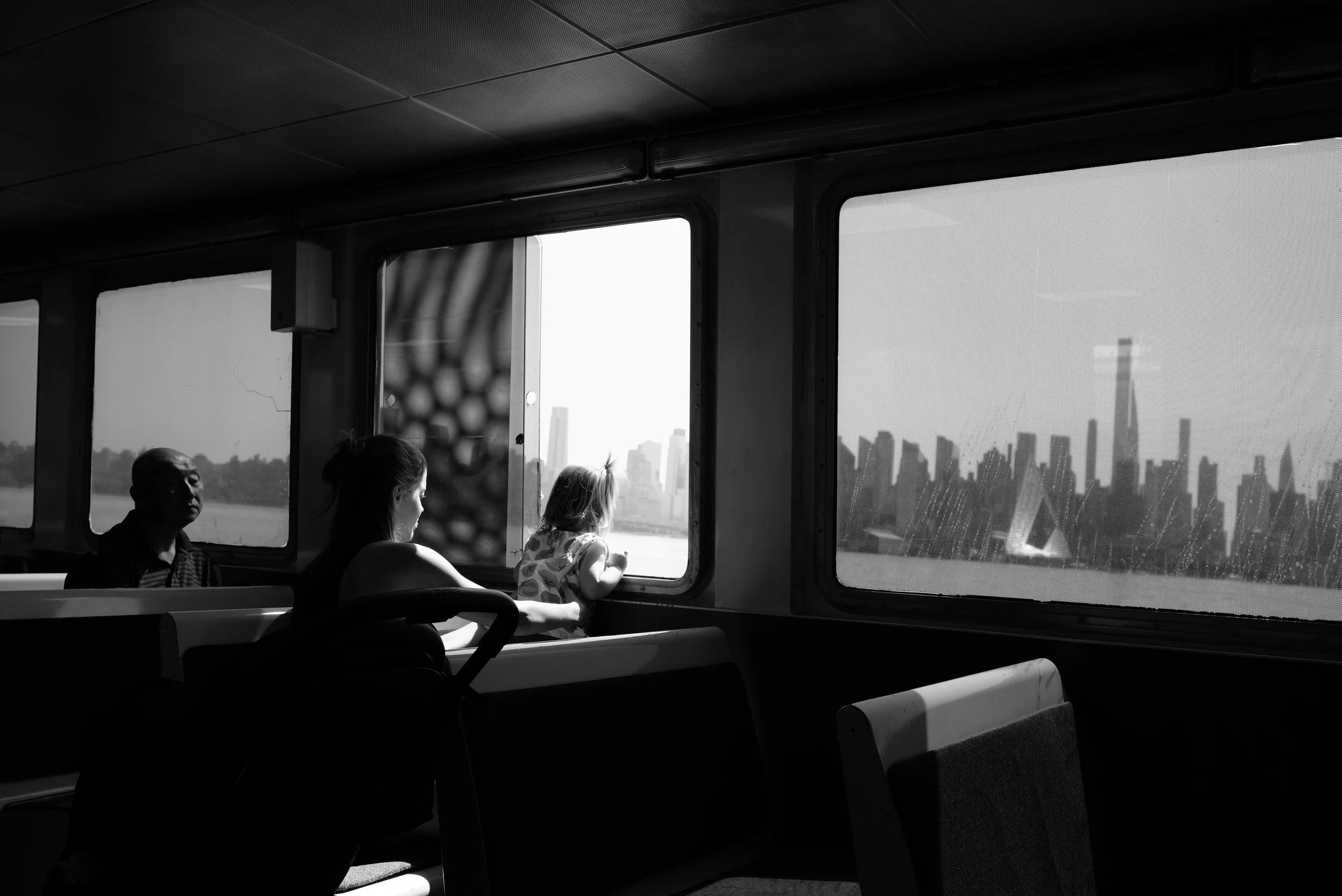 Black and white photo of three people looking out the windows of a ferry, with a city skyline in the background.