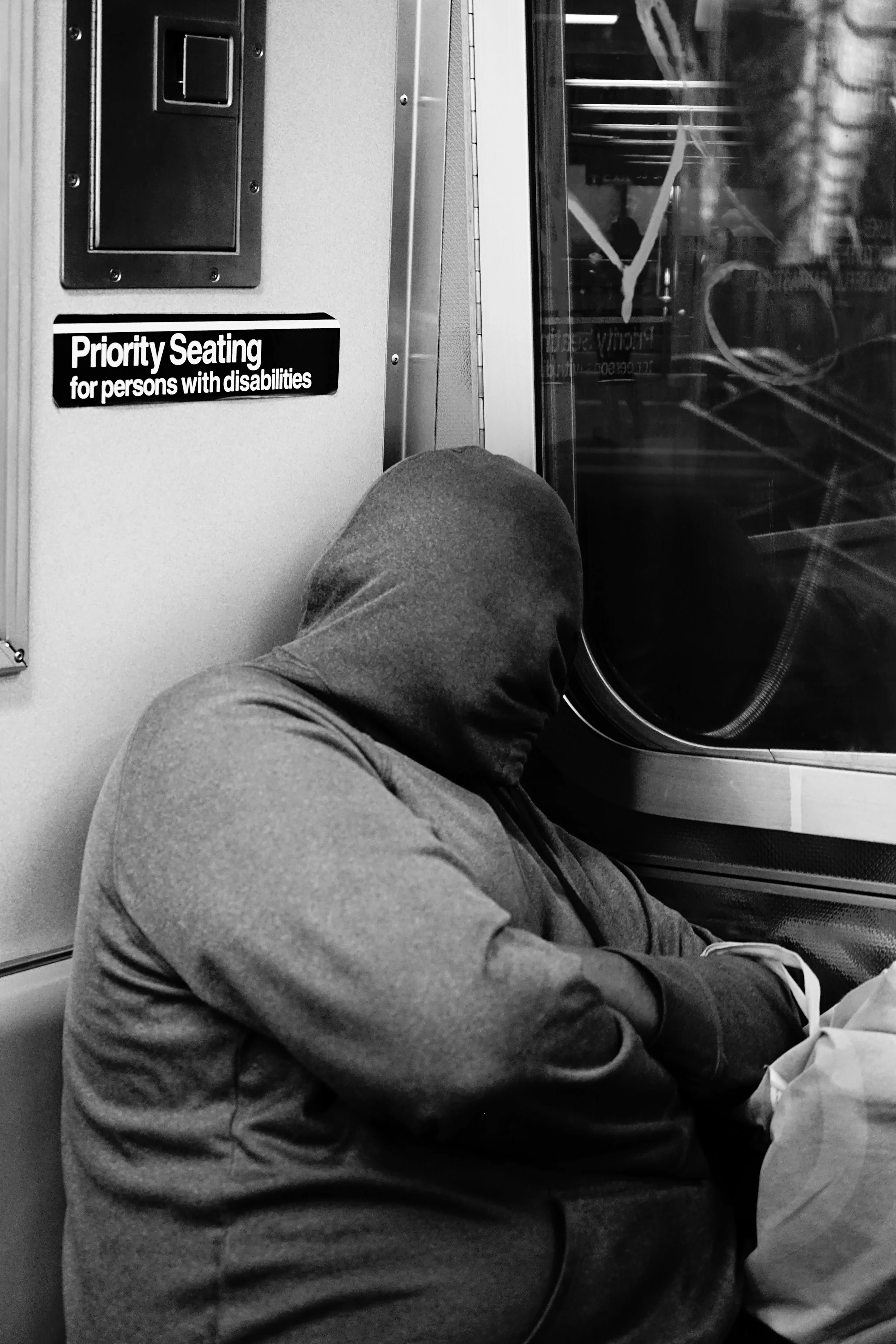 A person wearing a hoodie sleeping on a train seat next to a window, with a sign indicating priority seating for persons with disabilities.