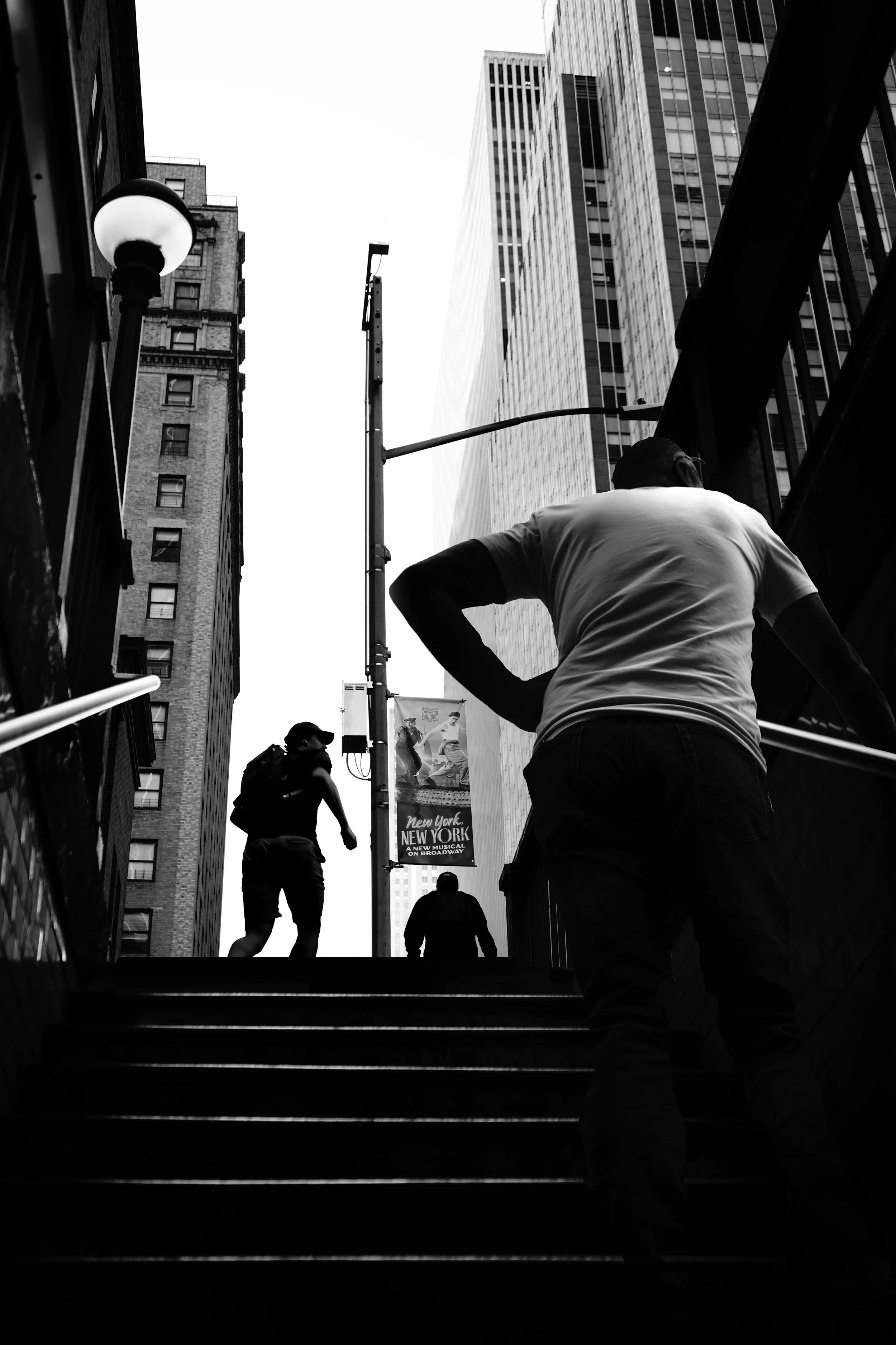 Silhouettes of people ascending stairs in a city with tall buildings and a billboard promoting a Broadway musical.