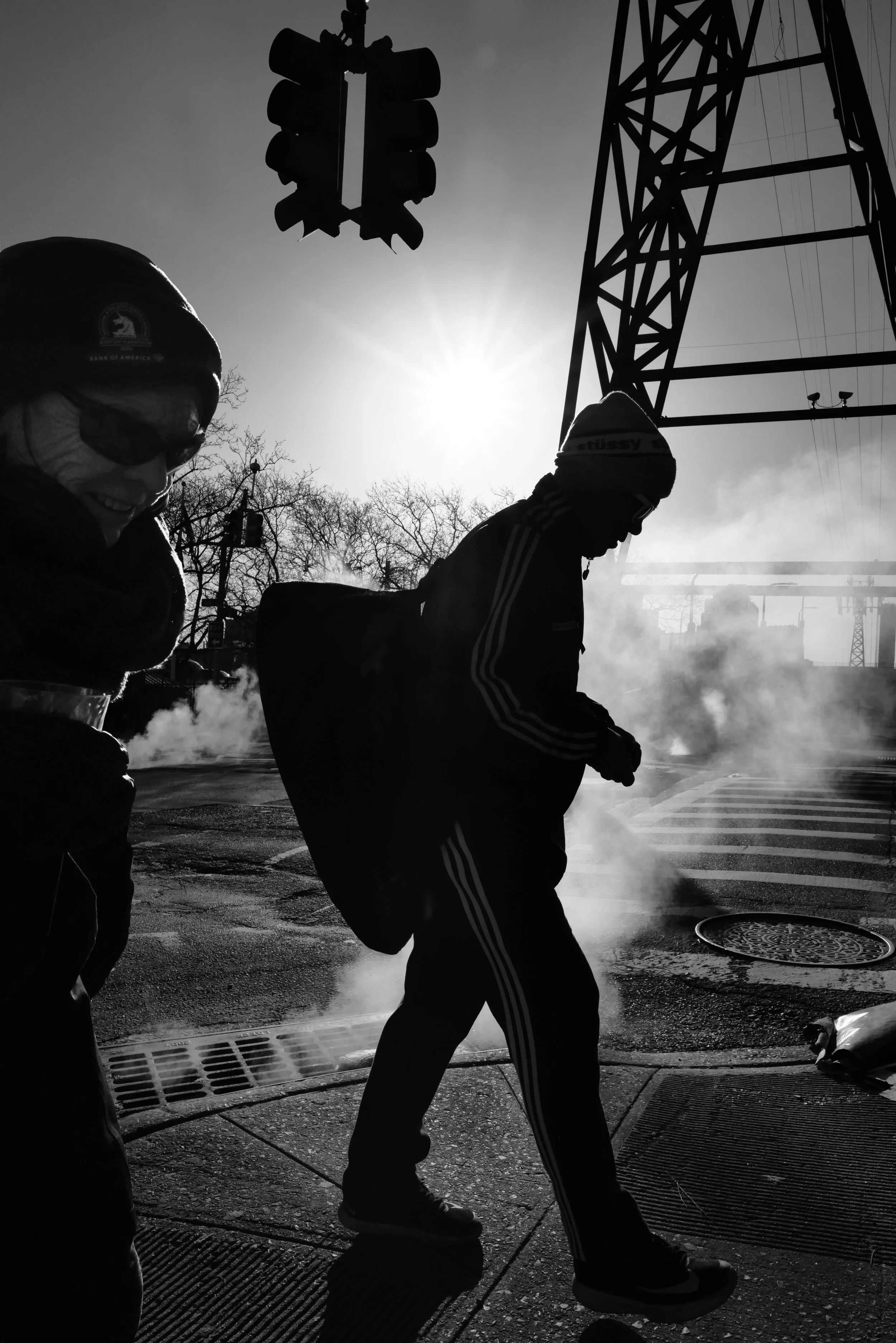 Black and white photo of two people walking on a street with steam rising from the ground. One person on the left is smiling and wearing glasses and a hat, while the person on the right is wearing a hooded jacket and a beanie. The sun is shining brightly behind them, with a large tower structure and traffic light above.