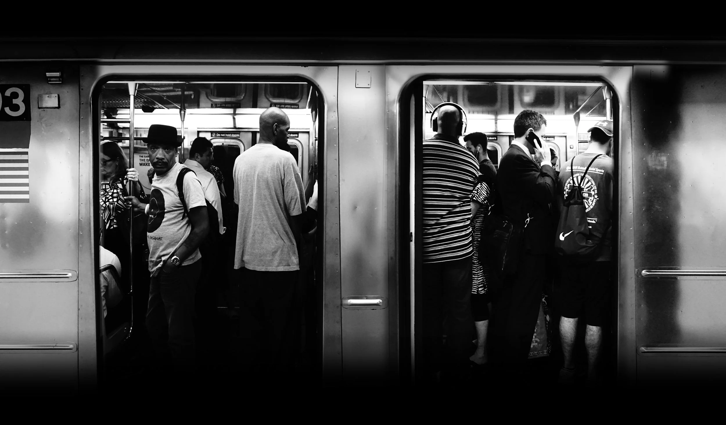 Black and white photo of people standing and waiting on a subway platform, seen through open subway train doors.