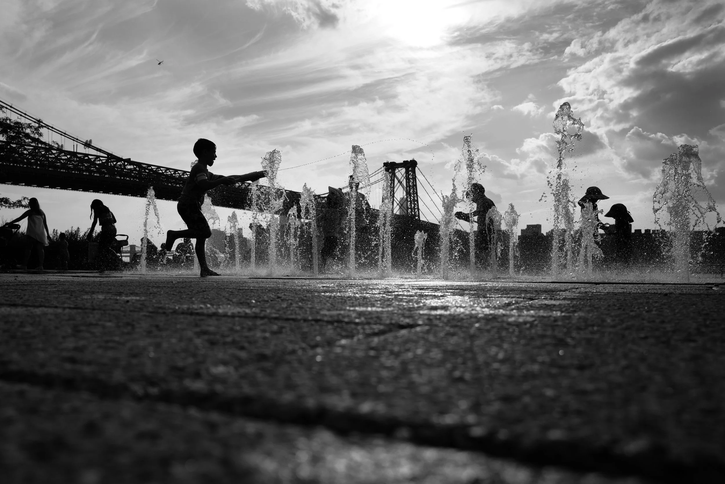 Silhouettes of children playing in a splash park with water fountains, bridge in the background, under a partly cloudy sky.