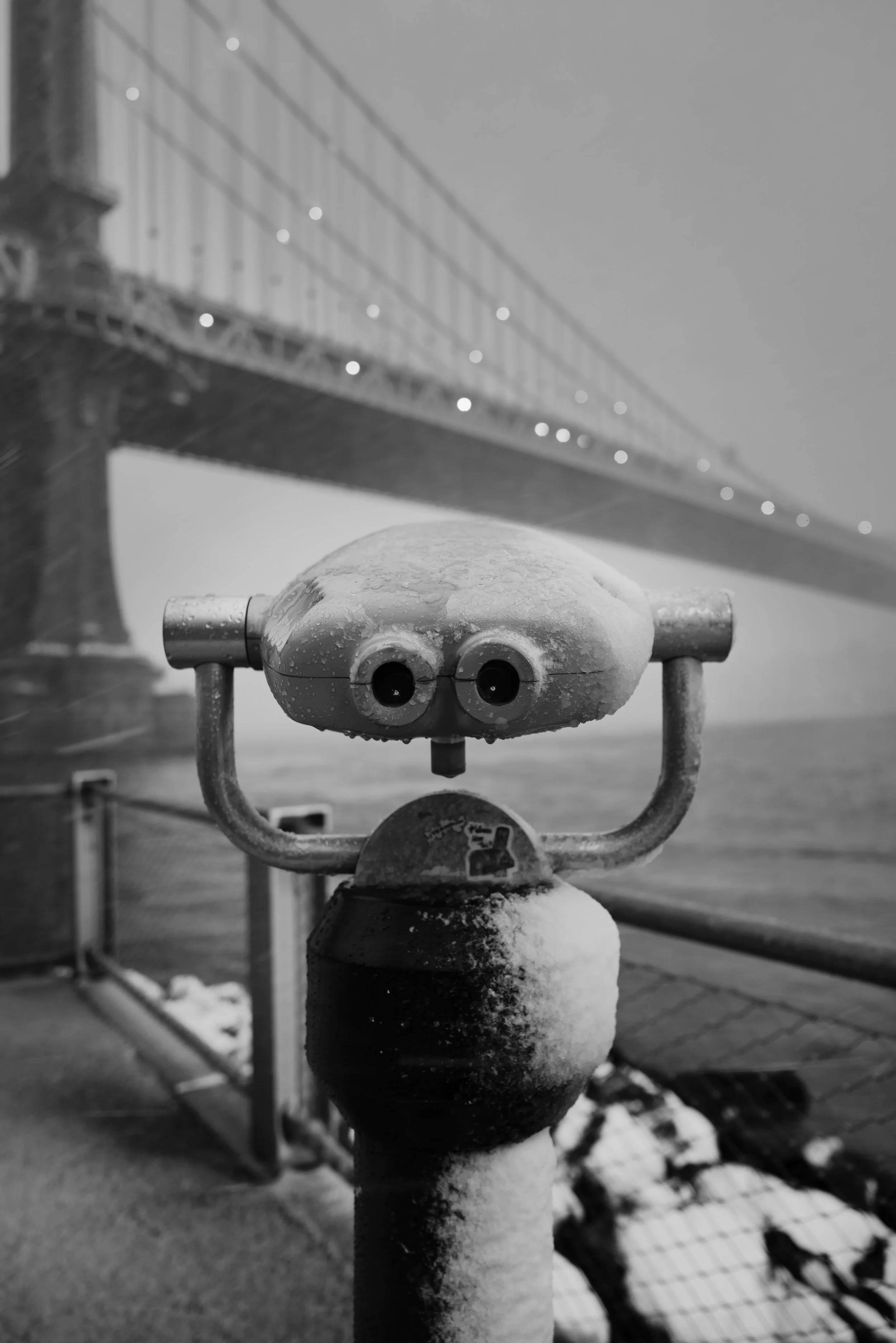 A snow-covered coin-operated binocular viewer on a pier with the Brooklyn Bridge in the background, foggy weather, black and white photo.