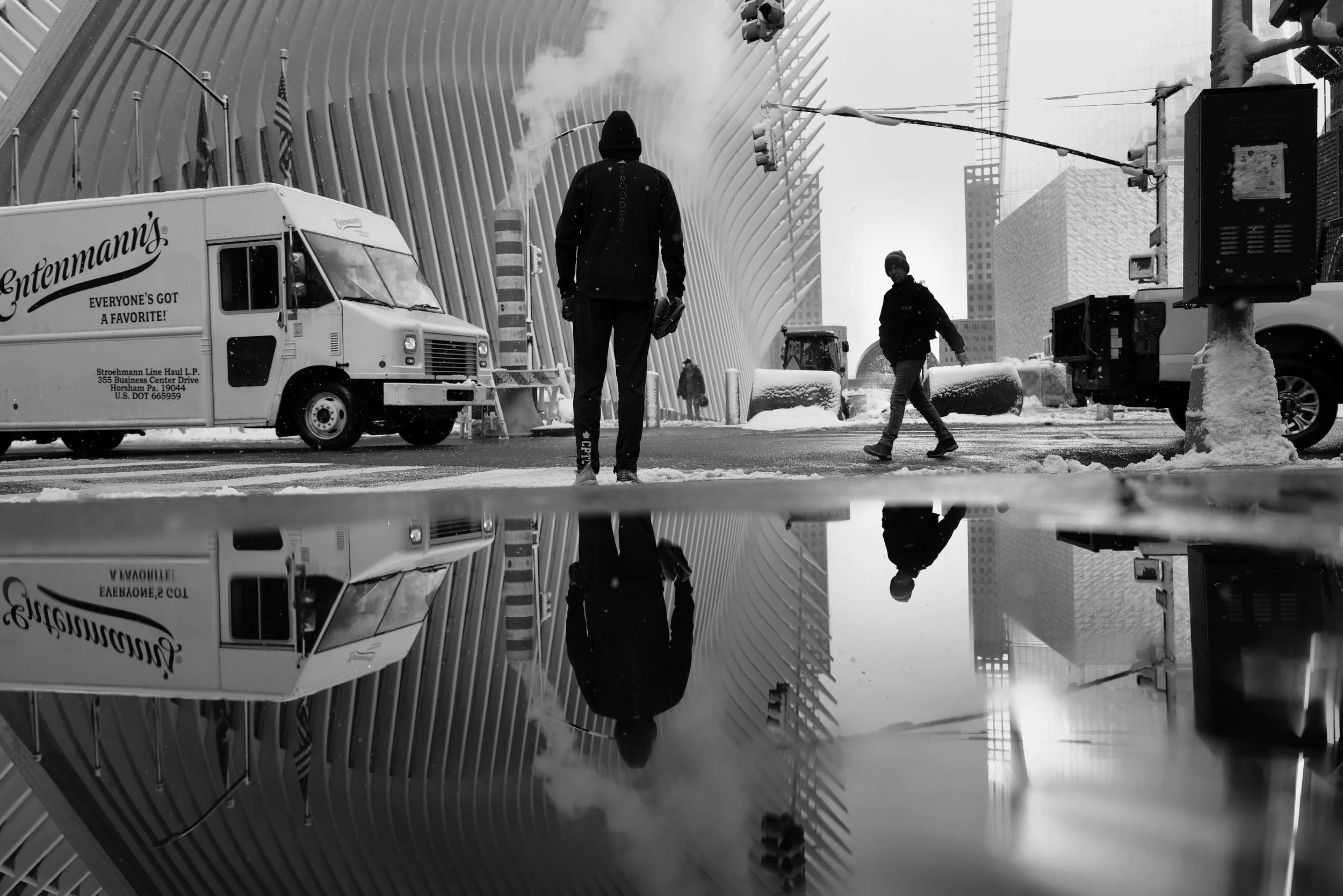 Black and white photo of people walking on a snowy city street, with a reflection of the scene on a puddle. There are snowplows, a delivery truck, and modern architectural structures in the background.