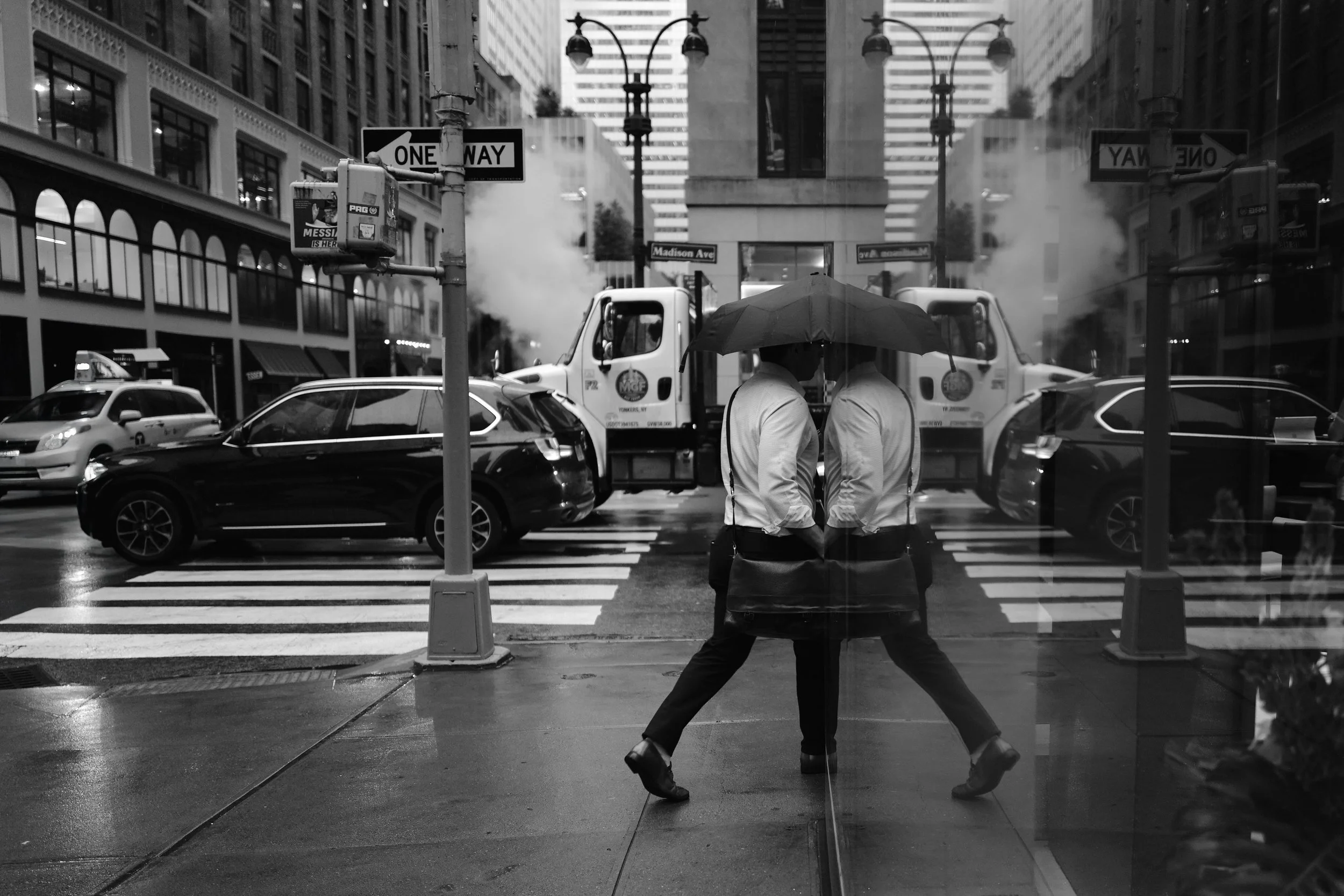 Two people with umbrellas walking on a city sidewalk, with their reflection visible on a glass window, and traffic including cars and a steam-venting street cleaning machine in the background.