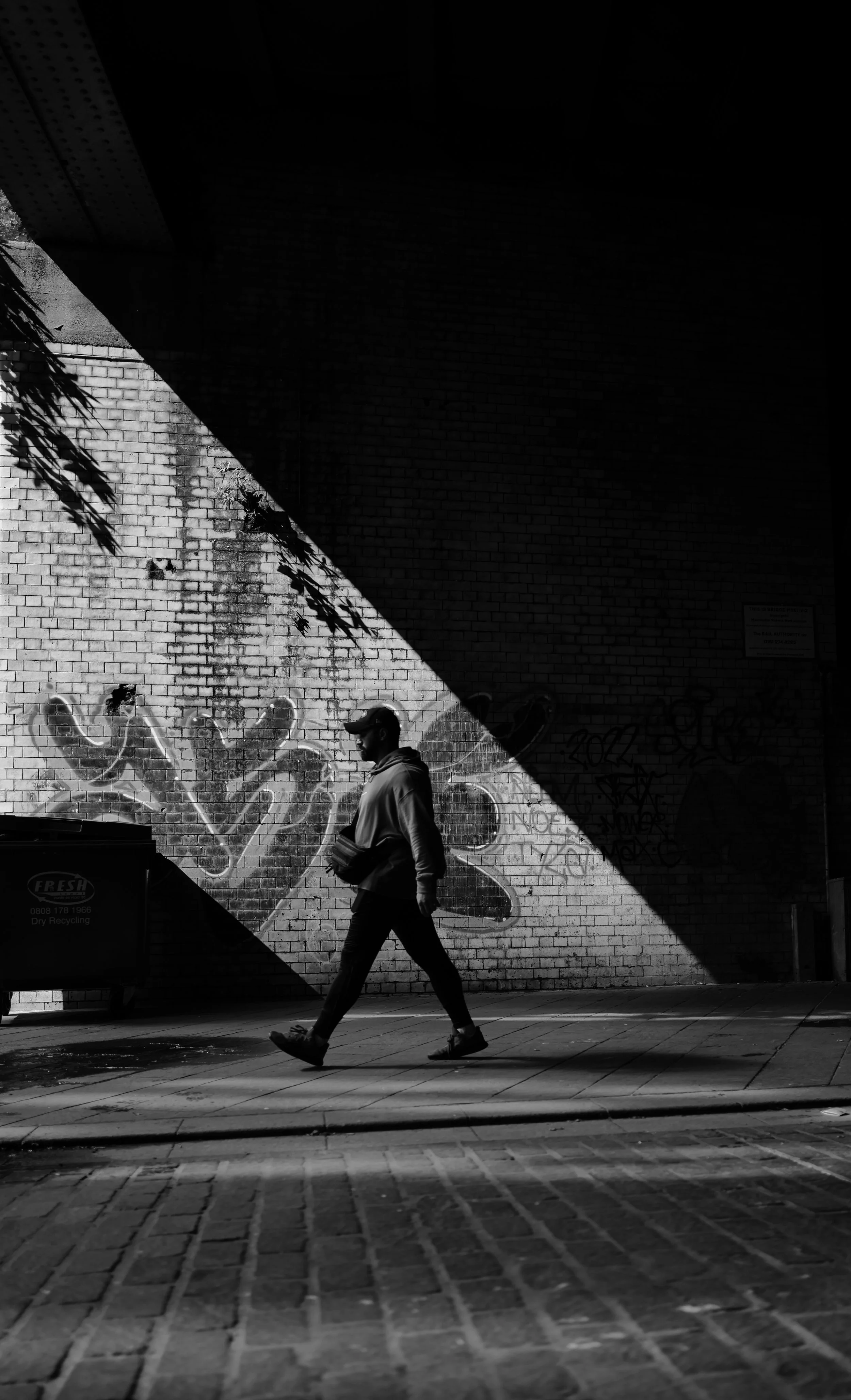 A person walking on a city sidewalk in front of a graffiti-covered brick wall, with shadows and sunlight creating a stark contrast.