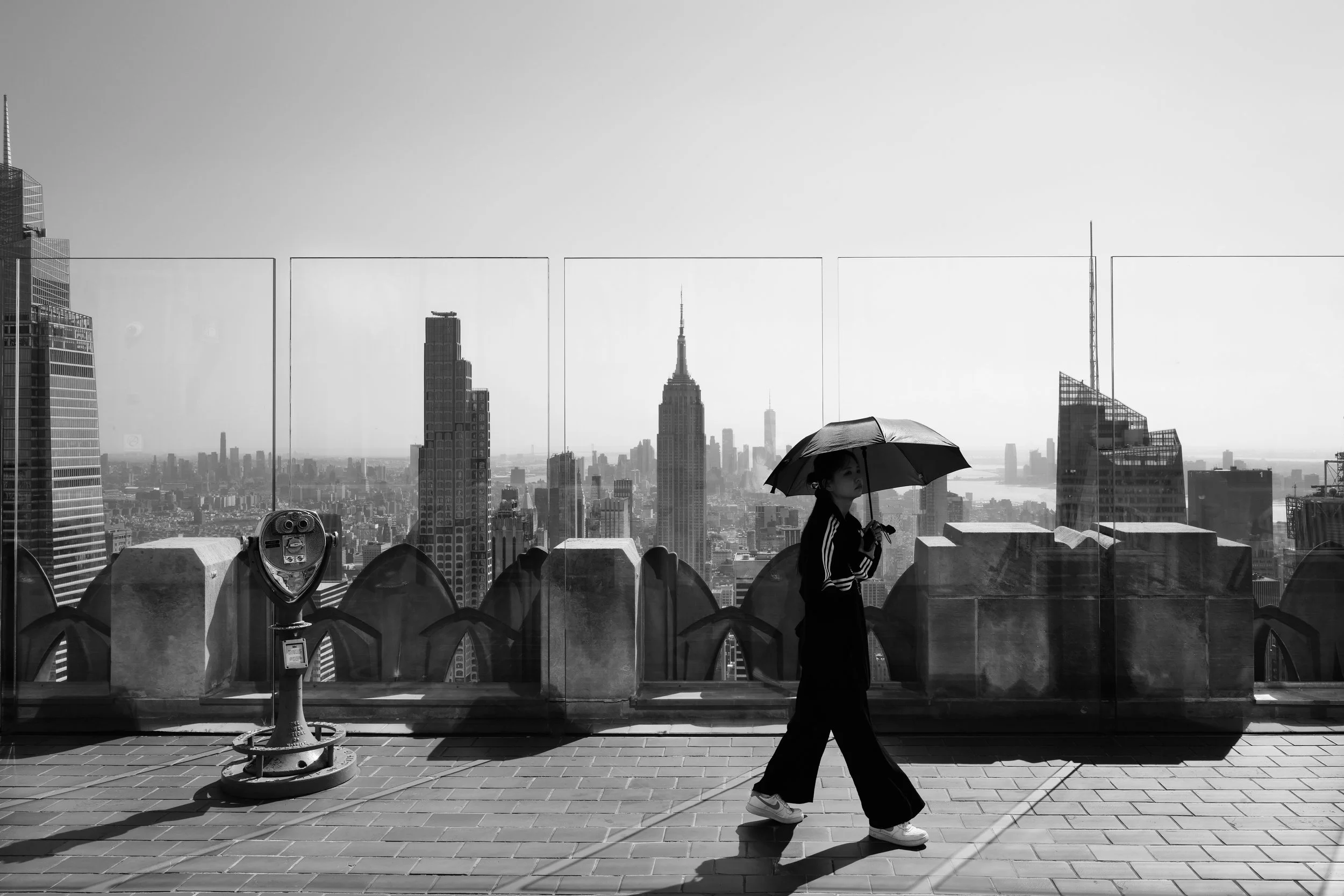 A woman walking with an umbrella on a rooftop with New York City skyline, including the Empire State Building, in the background.