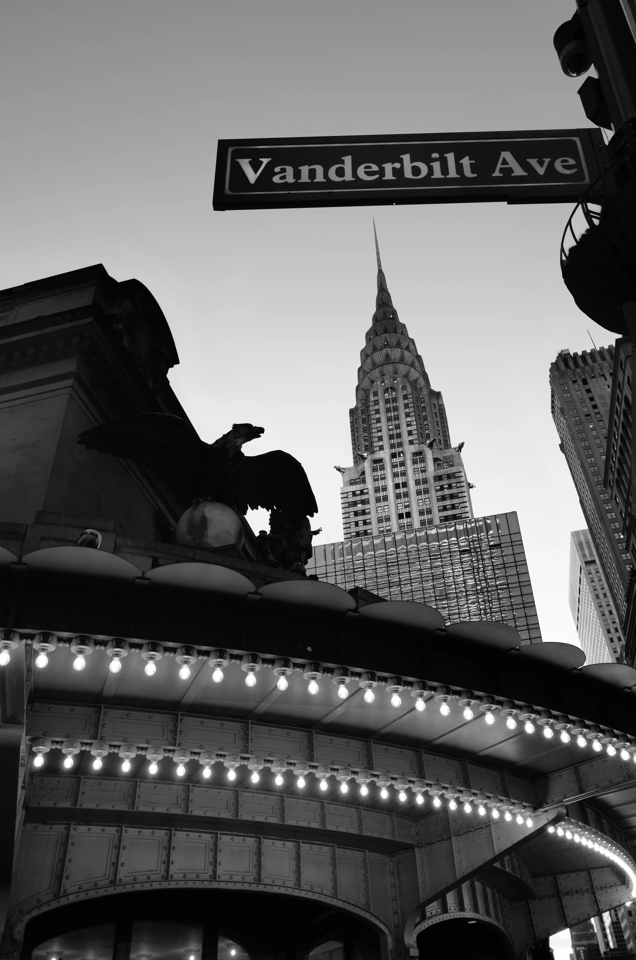 Black and white photo of the Empire State Building in New York City with a street sign for Vanderbilt Avenue in the foreground, along with a decorative eagle sculpture and string lights.