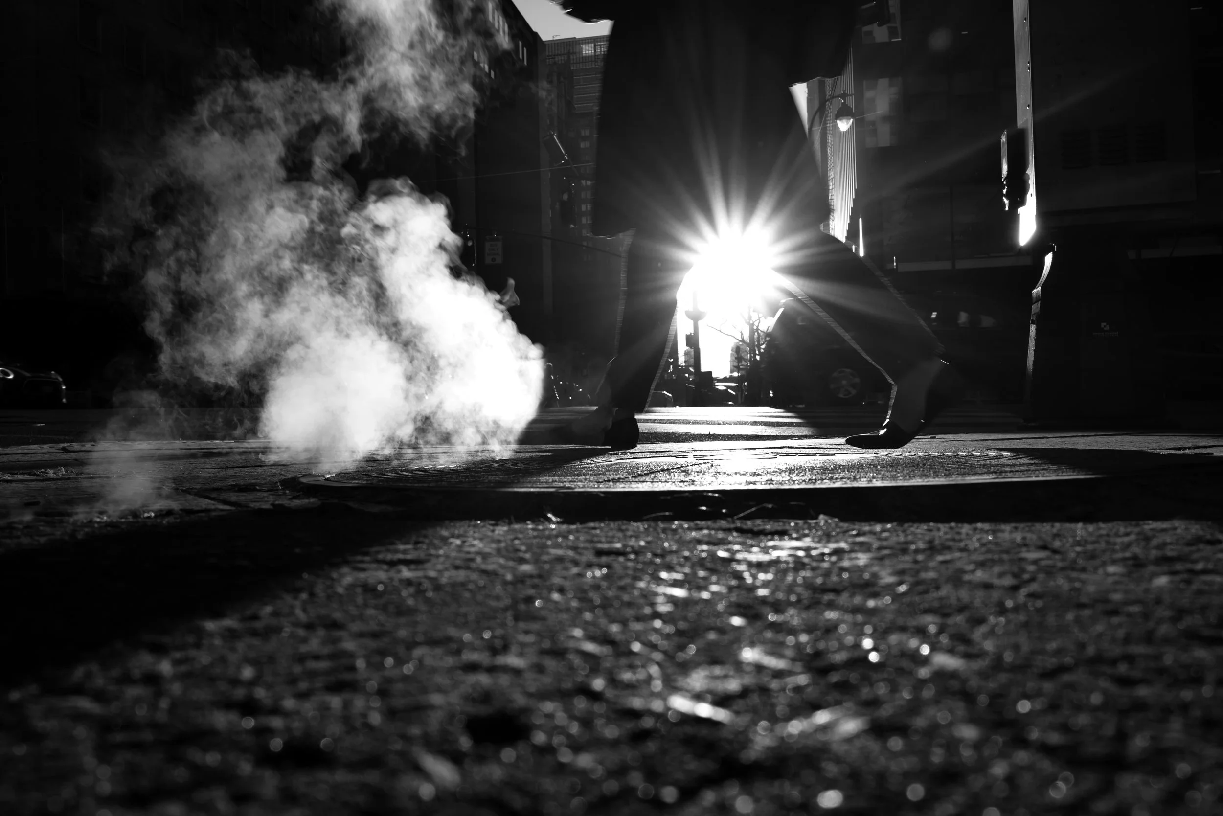 A person bending over on a city street with steam rising from the ground, backlit by a bright sun.