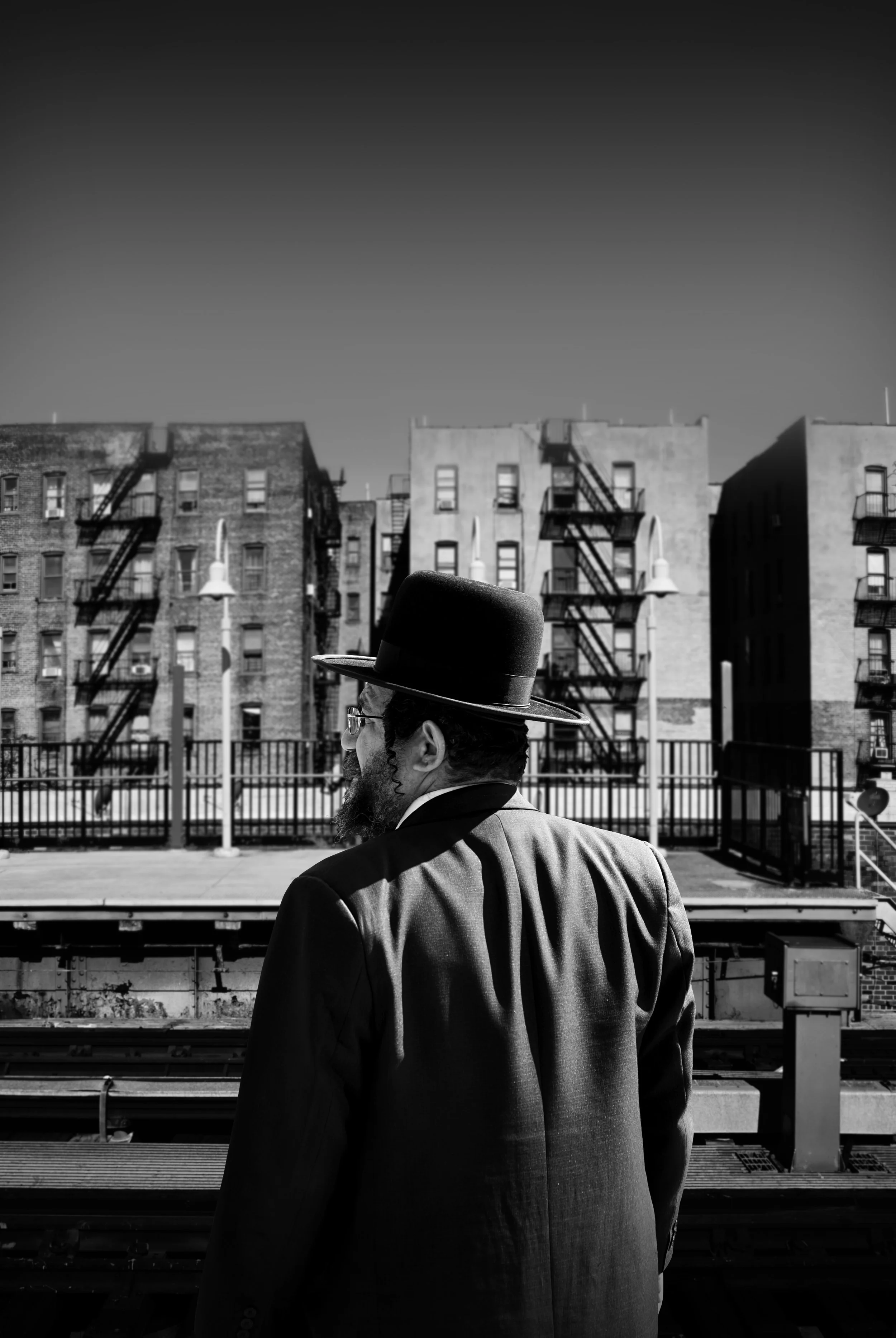 A man wearing a hat and suit standing near a train track in an urban environment, with apartment buildings and fire escapes in the background, shown in black and white.