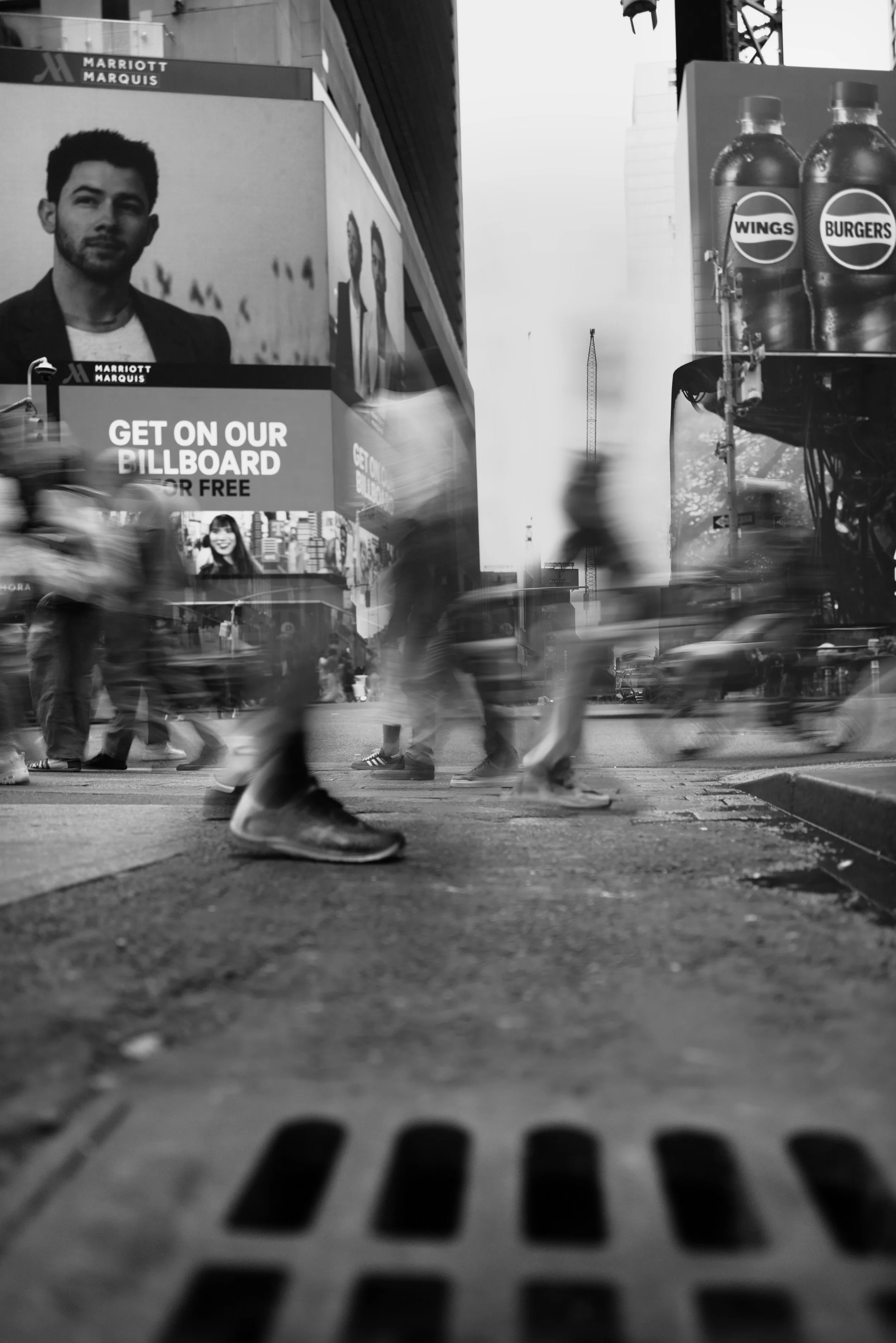 Black and white photo of a busy city street with blurred pedestrians walking past. In the background, there are large billboards including one advertising WINGS and BURGERS. The photo is taken from a low angle, focusing on the sidewalk and the movement of the crowd.