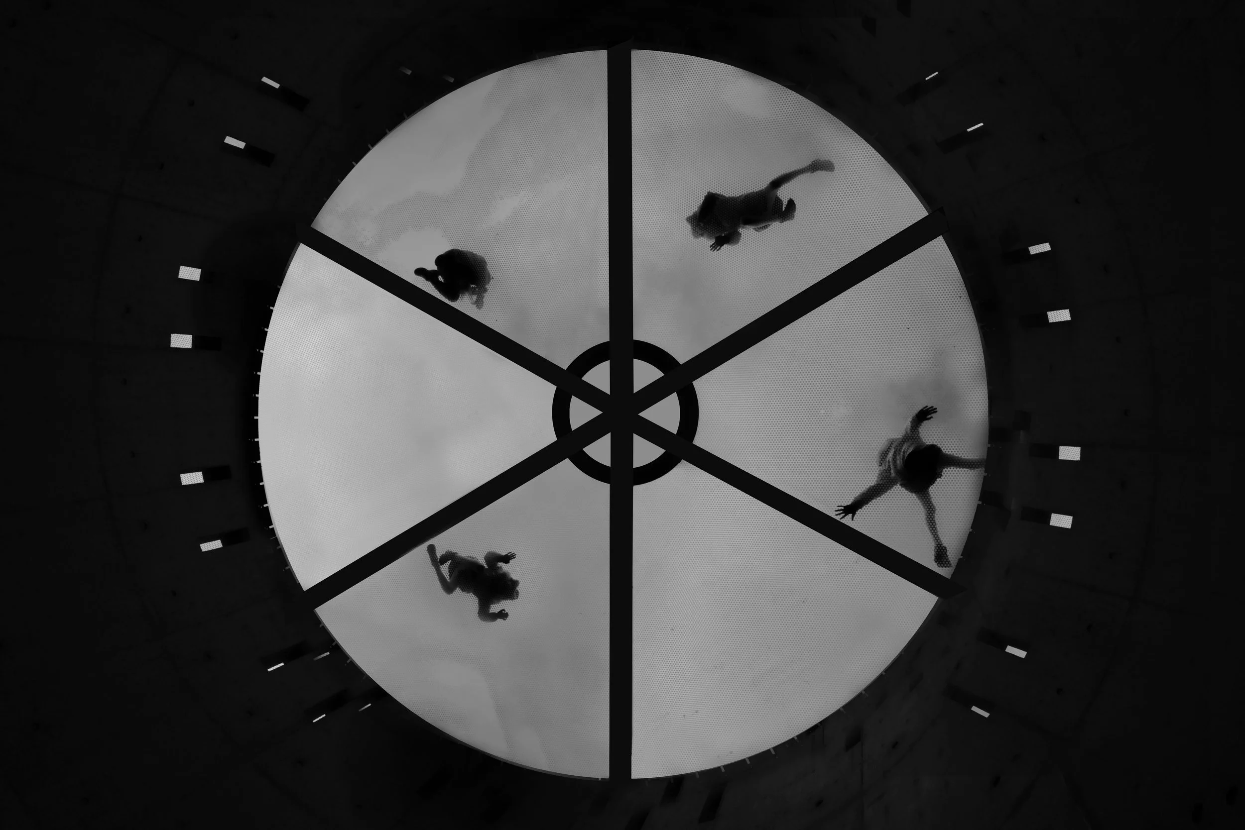 People walking on a circular glass floor with spider web design, looking upward through the glass at their reflections.
