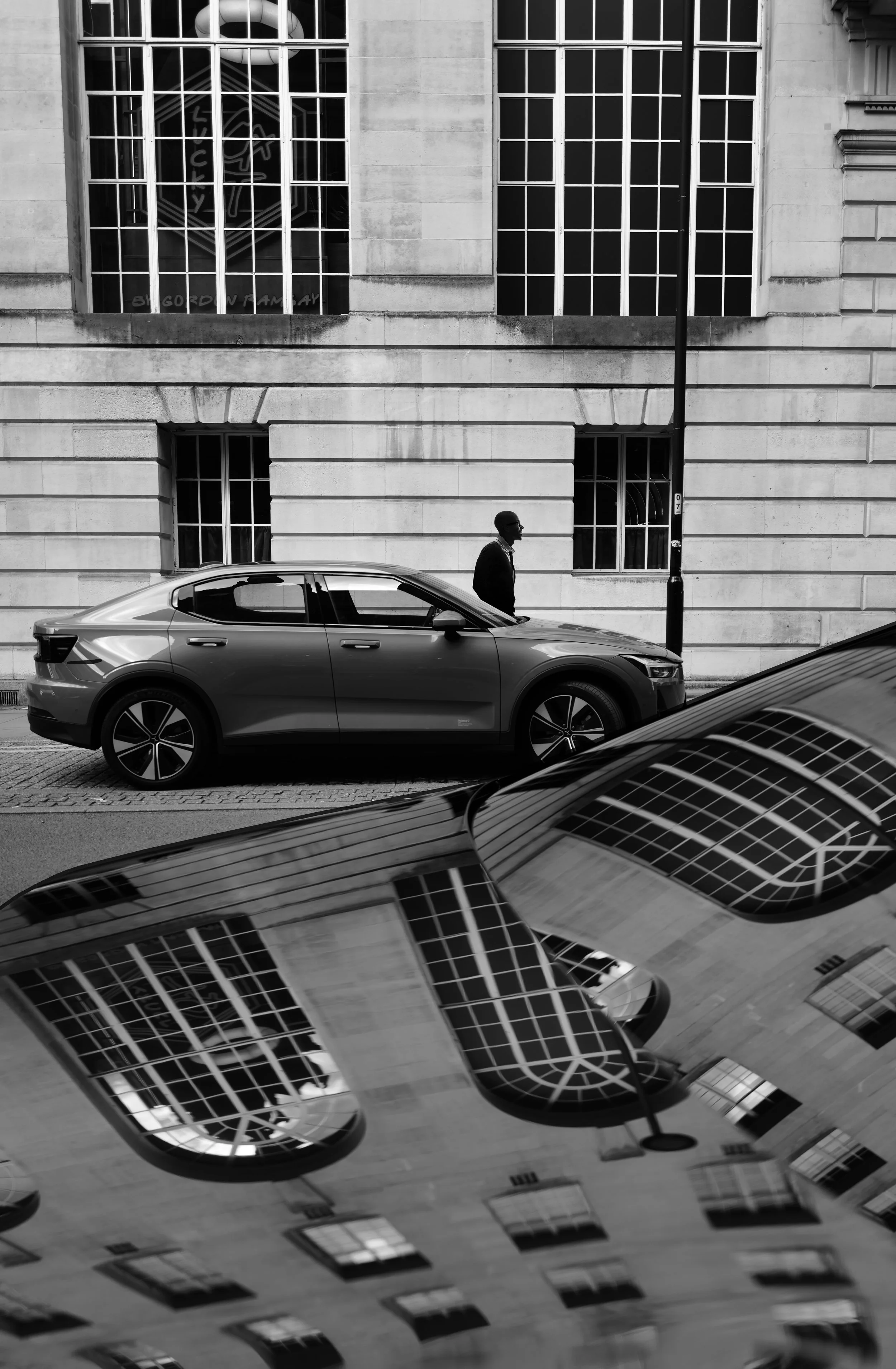 A man walking on the sidewalk near parked cars in front of a building with large windows, one reflecting the street scene, in black and white.