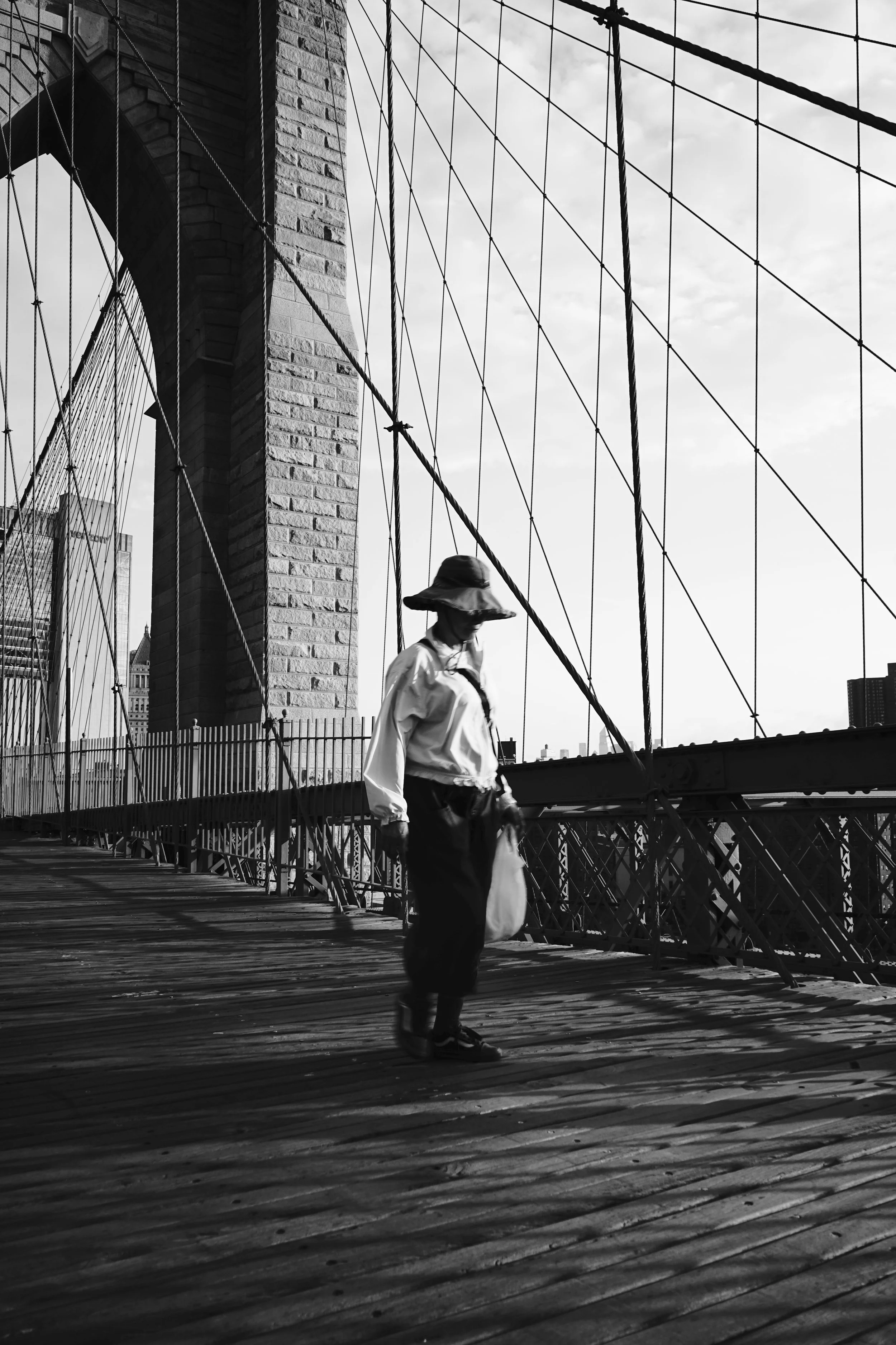 A woman walking on the Brooklyn Bridge in New York City during daytime, wearing a wide-brimmed hat, light-colored shirt, dark pants, and sandals, carrying a bag. The bridge's cables and stone tower are visible in the background.