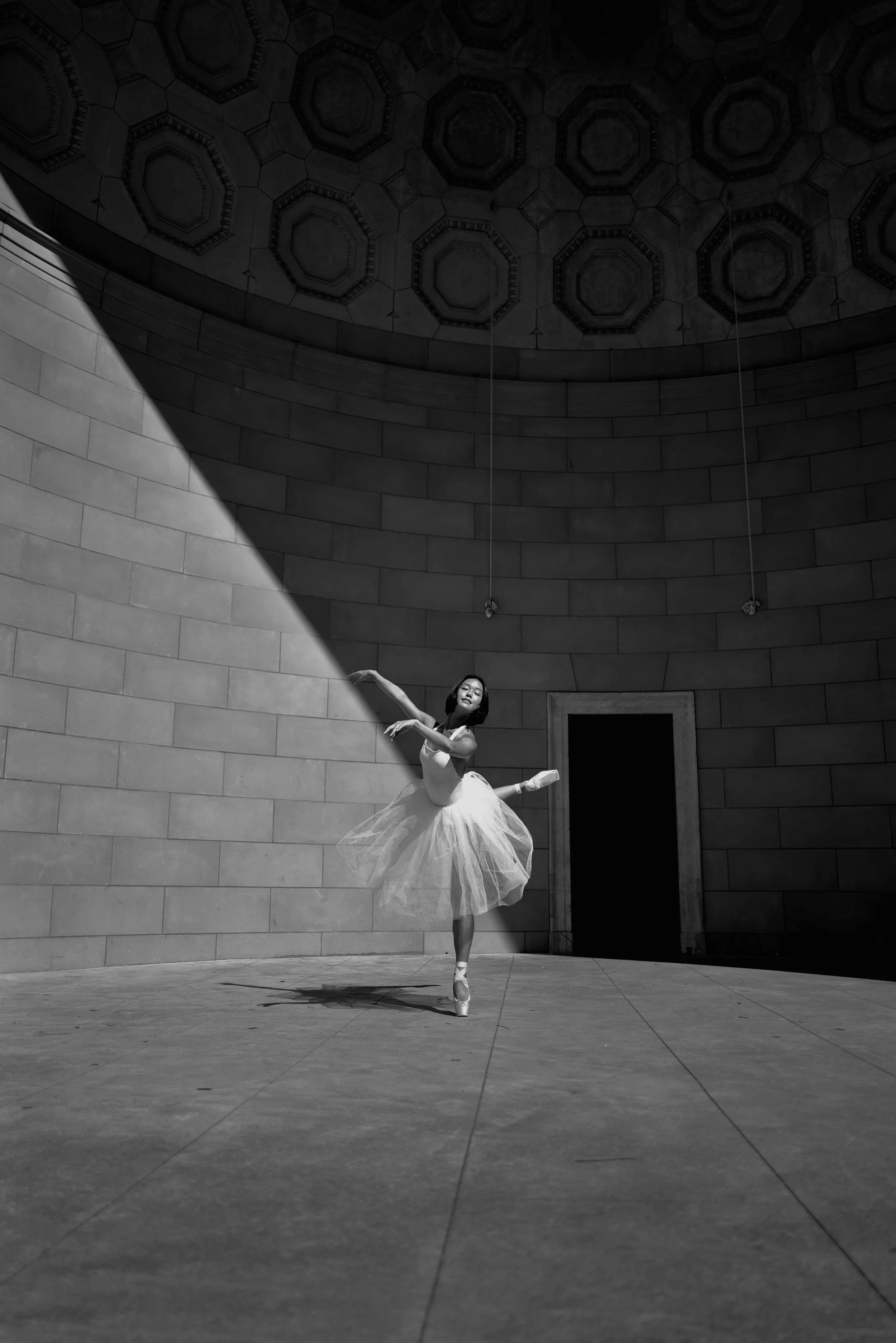 A ballerina in a tutu performing a dance move inside a large, circular, domed hall with decorative ceiling patterns in black and white.