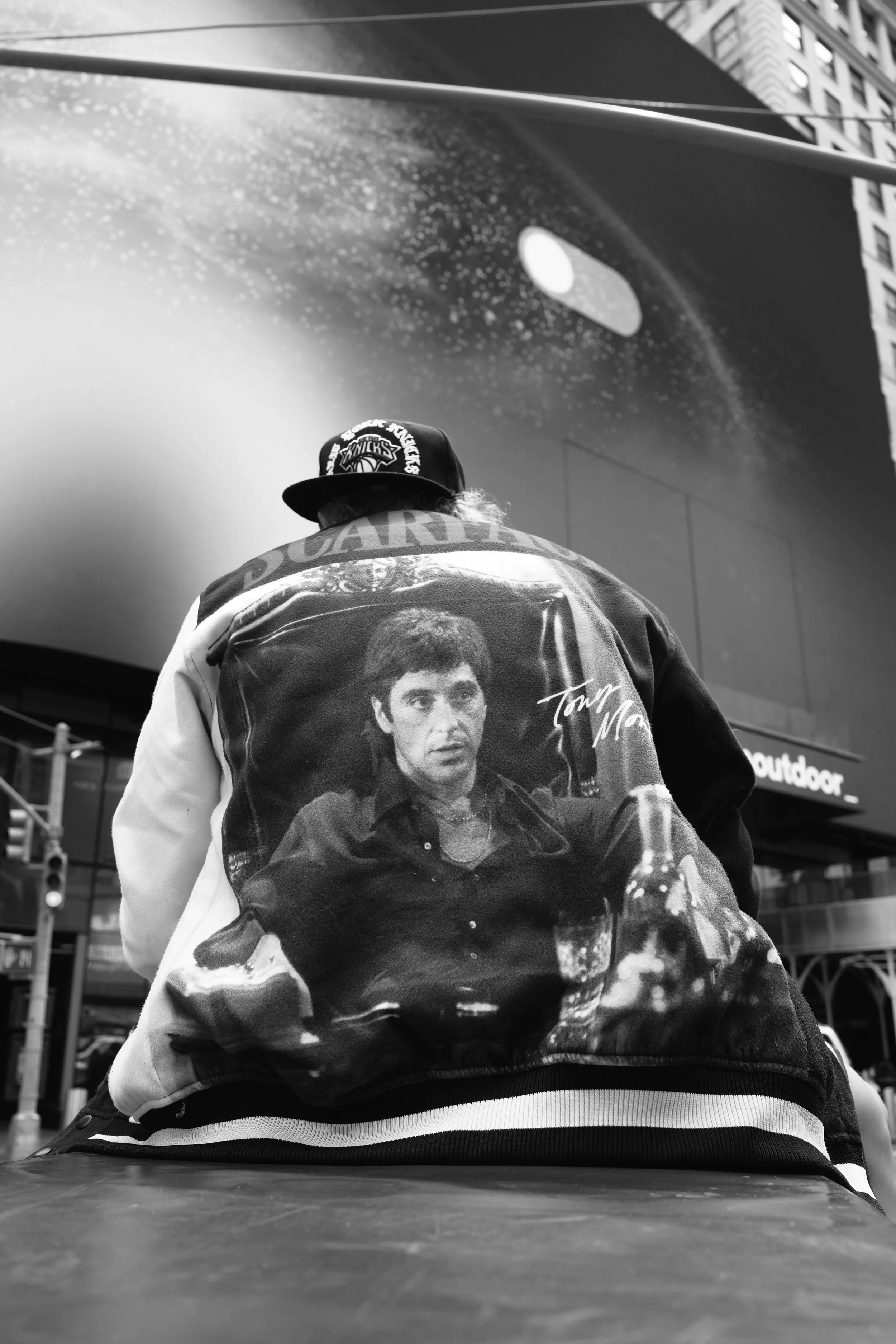Back of a person wearing a jacket with a large image of Tony Montana from Scarface, signature, and text. The person also wears a black cap with a New York Knicks logo and is seated outside in an urban environment.