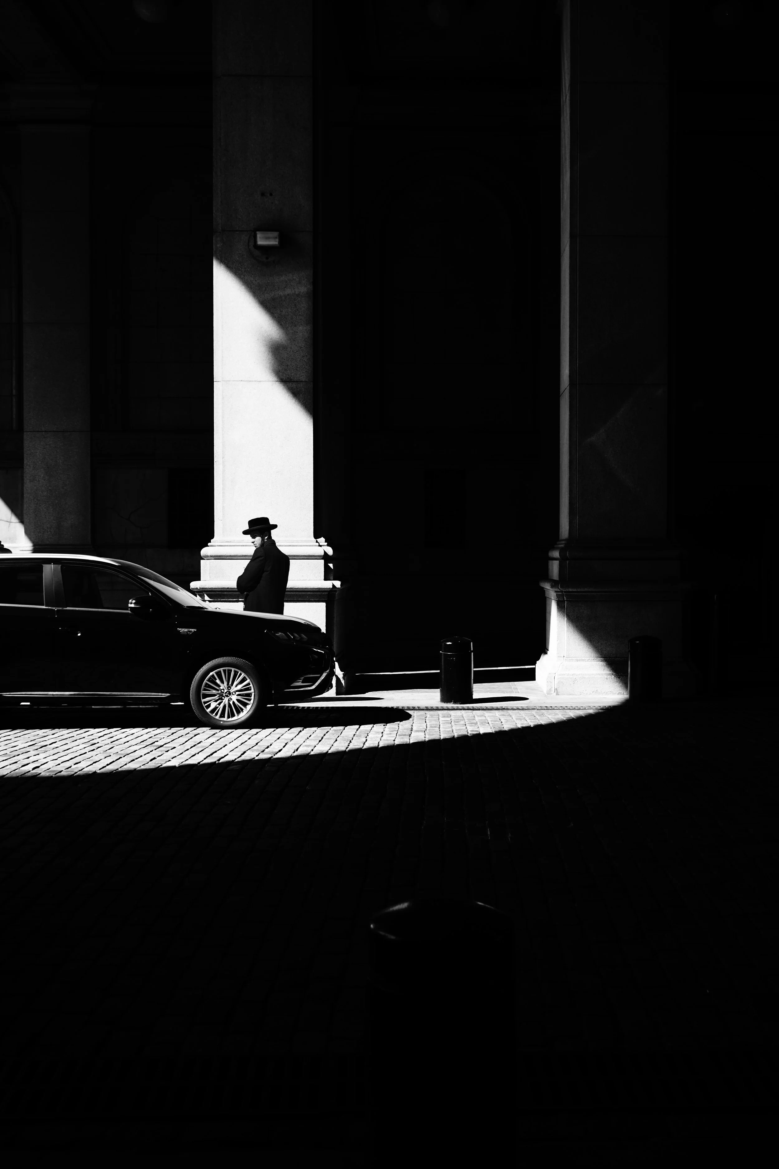A black and white photo of a person with a hat and coat standing outdoors next to a parked car with a large building in the background, with stark light and shadow patterns.
