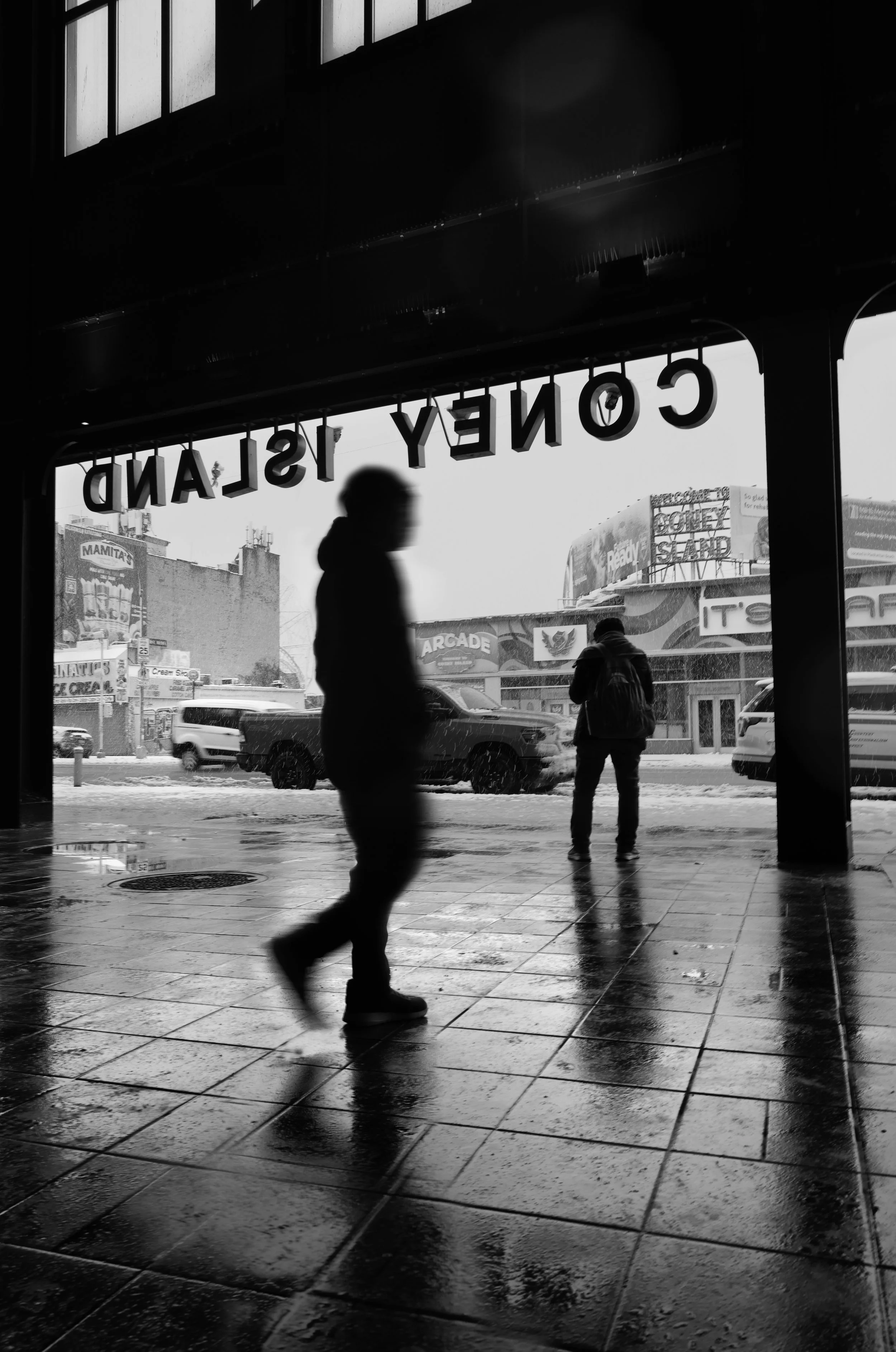 A black and white photo taken from inside a building, looking out through an entrance onto a street during snowy weather. Two people with backpacks are walking, and cars are visible on the street. The scene has wet, reflective pavement and signage, including a sign that partially reads 'CONELY ISLAND'.